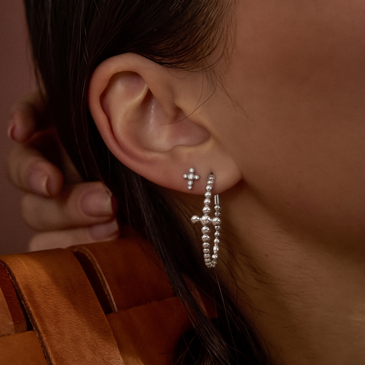 A close-up of a person's ear wearing two silver cross-shaped earrings, one small stud cross and one larger beaded hoop-style cross earring, against a warm brown background.