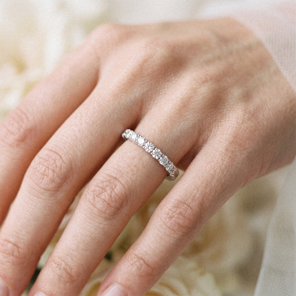A close-up of a hand wearing a delicate silver eternity ring set with small clear stones, photographed against a soft, blurred cream background.