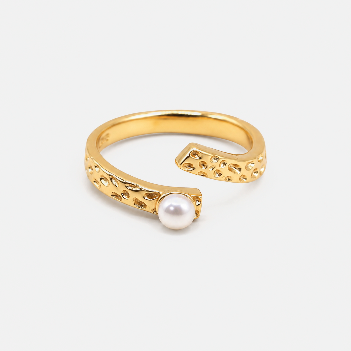 A gold open-bend ring featuring textured, hammered details and a single white pearl set in one of the ends, photographed on a clean, light gray background.