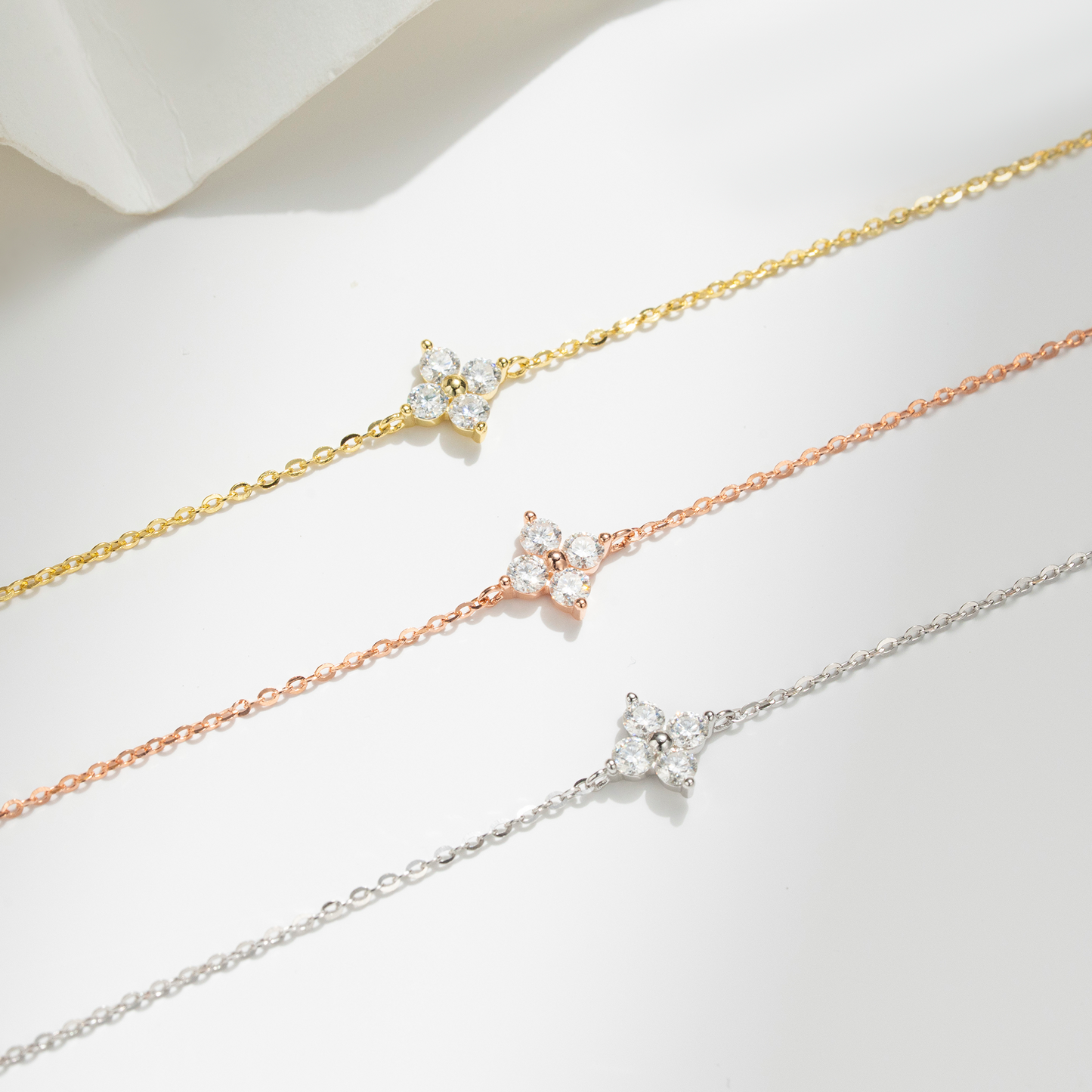 Three delicate chain bracelets in yellow gold, rose gold, and silver, each featuring a small flower-shaped cluster of clear cubic zirconia stones, arranged diagonally on a white surface.