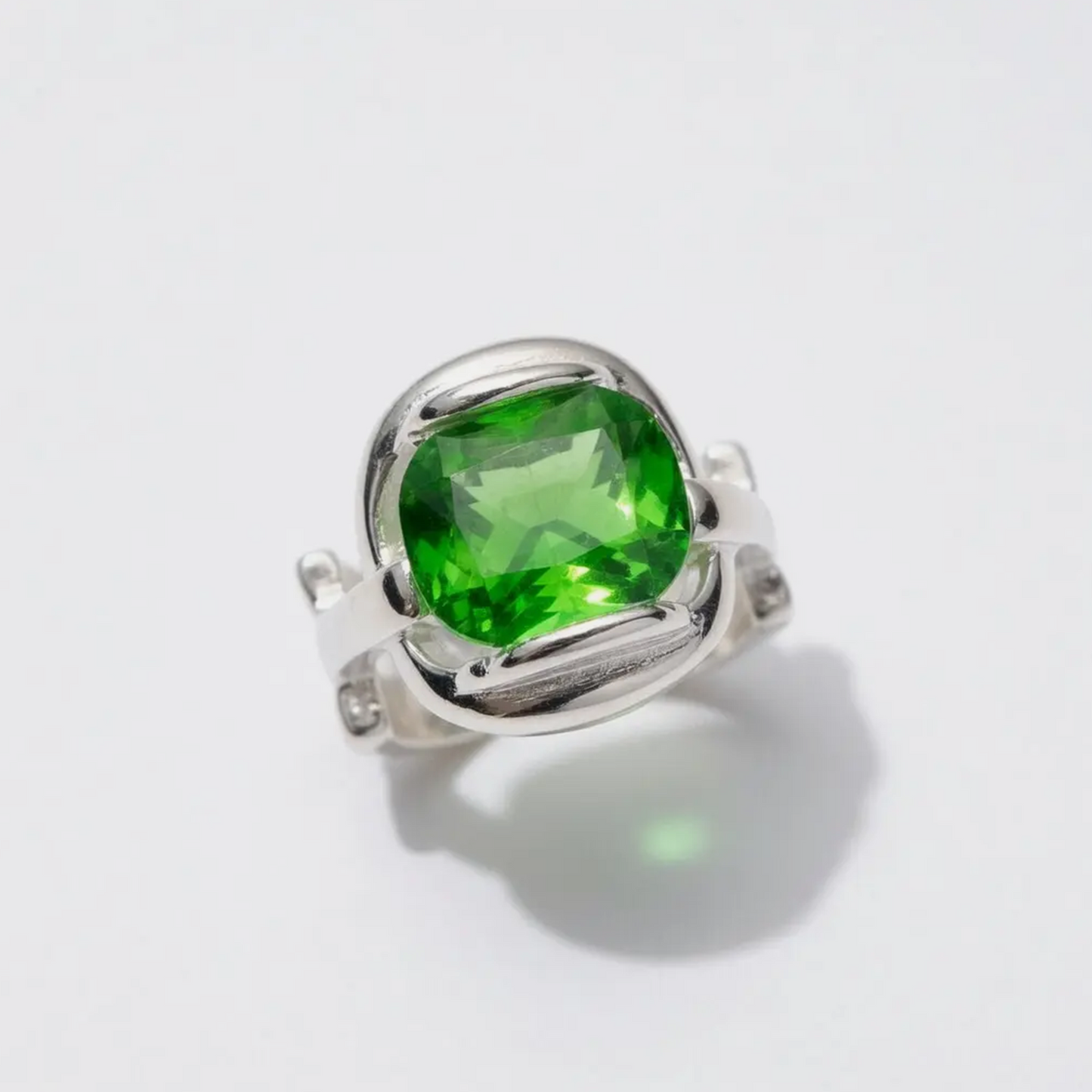 A silver ring featuring a large, cushion-cut green gemstone in a prong setting, photographed on a clean white background with a soft shadow.
