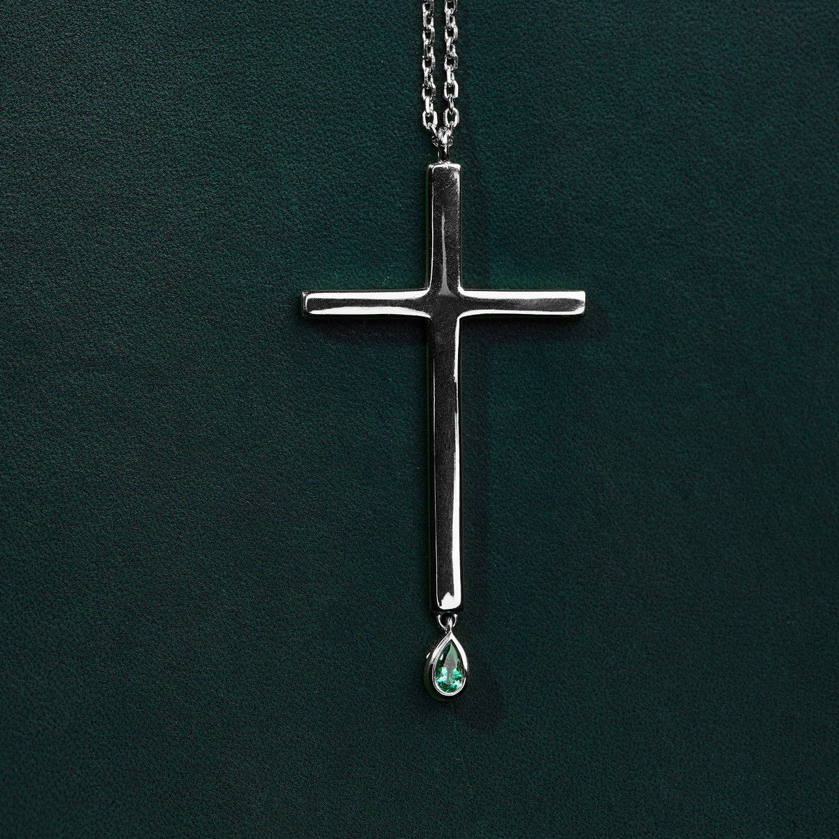 A silver cross pendant necklace with a small teardrop-shaped green gemstone hanging from the bottom, displayed against a dark green textured background.