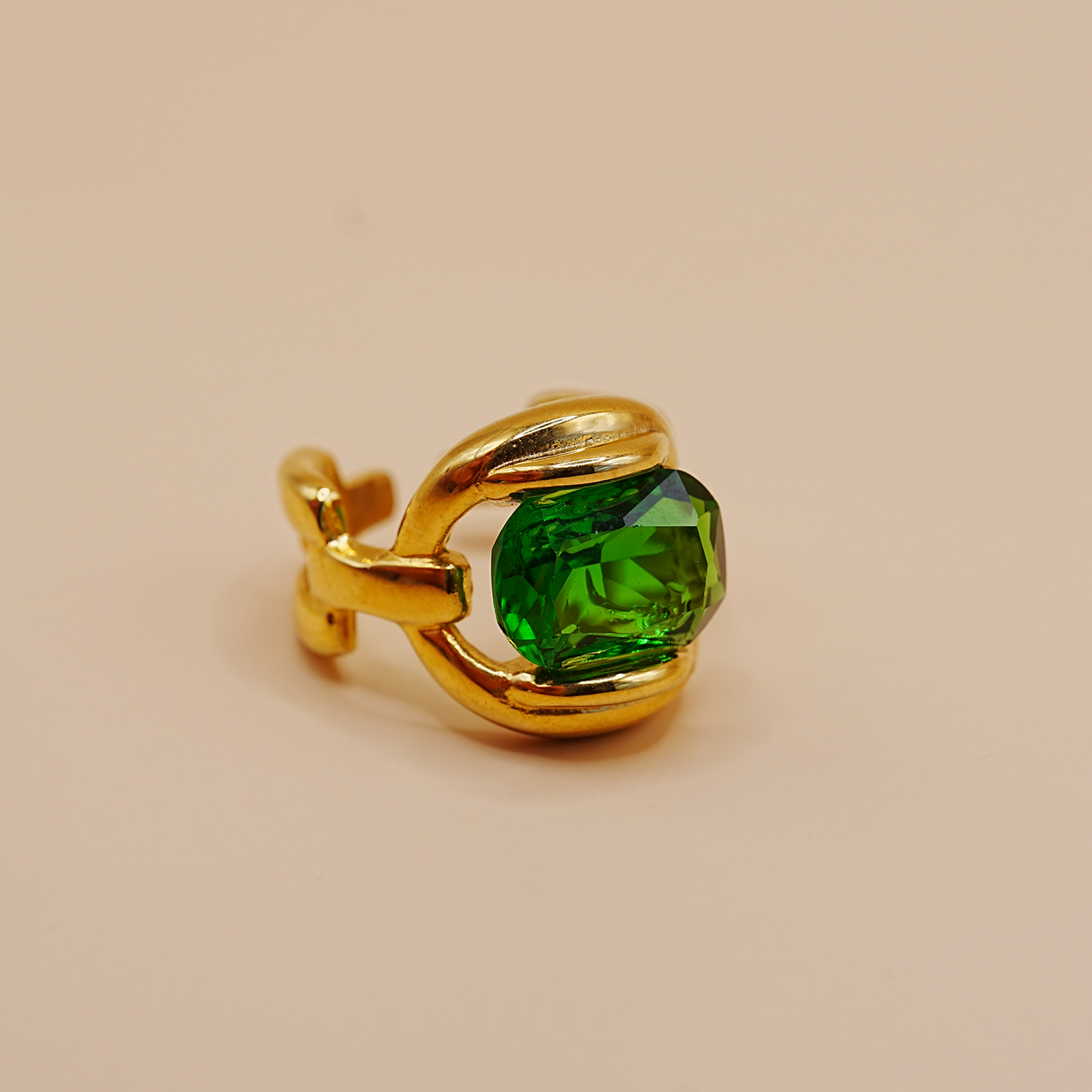 A gold adjustable ring featuring a large, faceted green gemstone, photographed on a solid beige background.