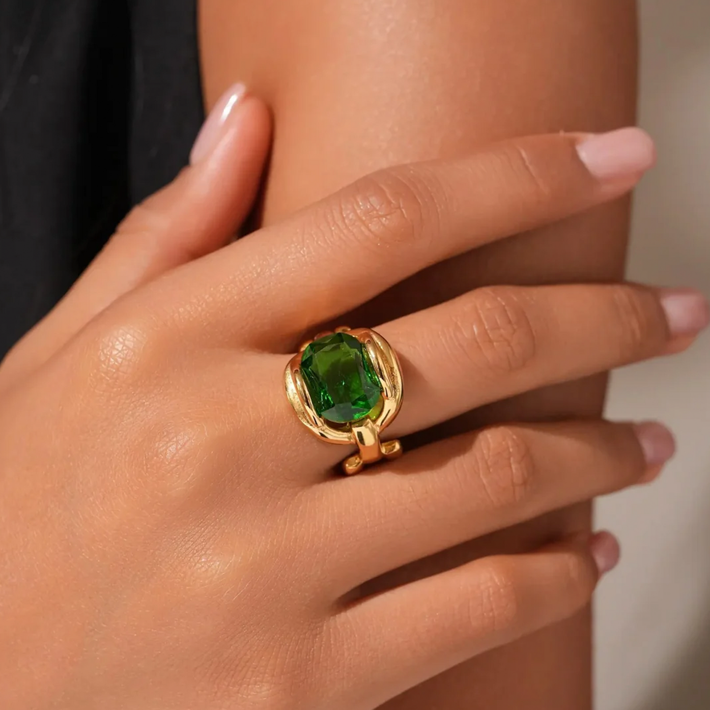 A close-up lifestyle shot of a gold ring with a large green cushion-cut gemstone worn on a person's finger.