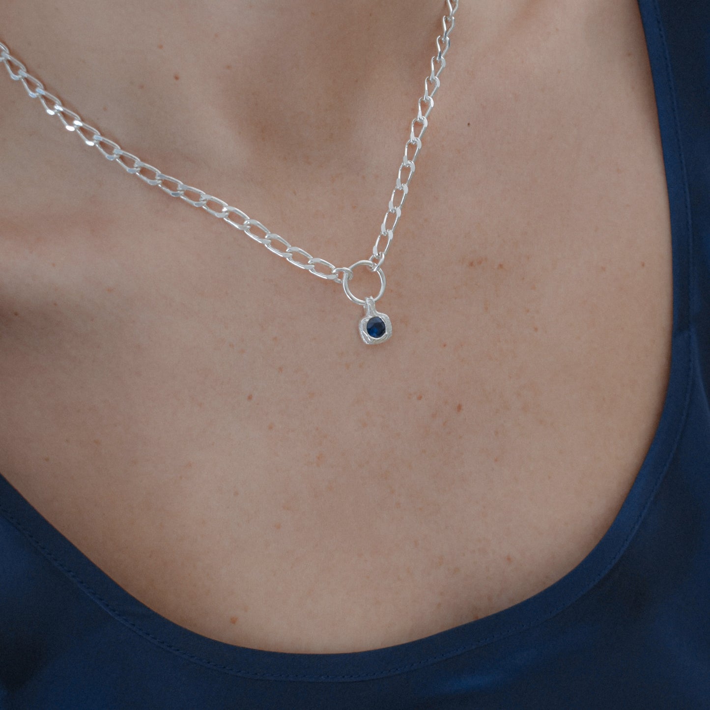 A close-up of a silver chain necklace with a small circular pendant featuring a dark blue stone, worn on a model's neck over a navy blue top.