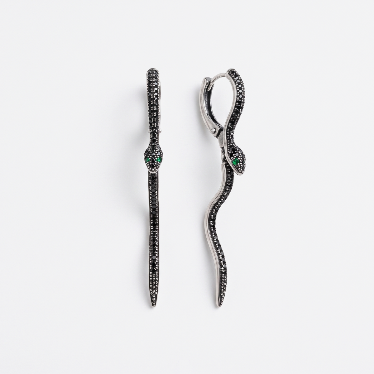 A pair of silver snake drop earrings paved with small black stones, featuring a small green stone accent at the head, shown on a light gray background.