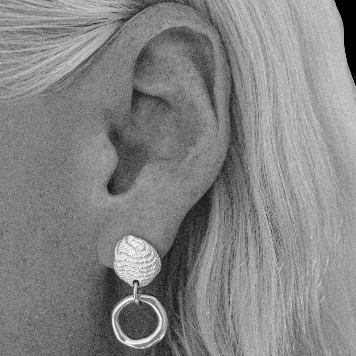 A close-up black and white photograph of a person's ear wearing a textured silver drop earring with a circular dangling element.