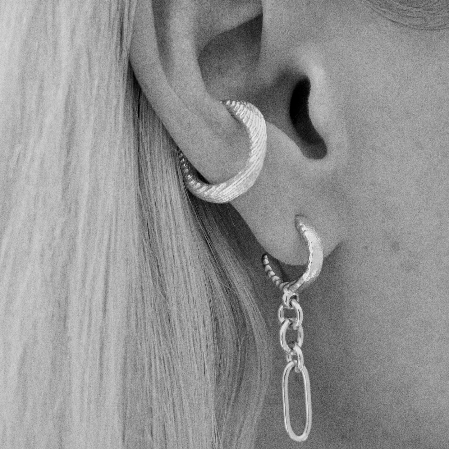 A close-up of a person's ear showcasing three silver-toned textured hoop earrings of varying sizes, including a larger cartilage hoop and two smaller lobe hoops with a dangling link charm.