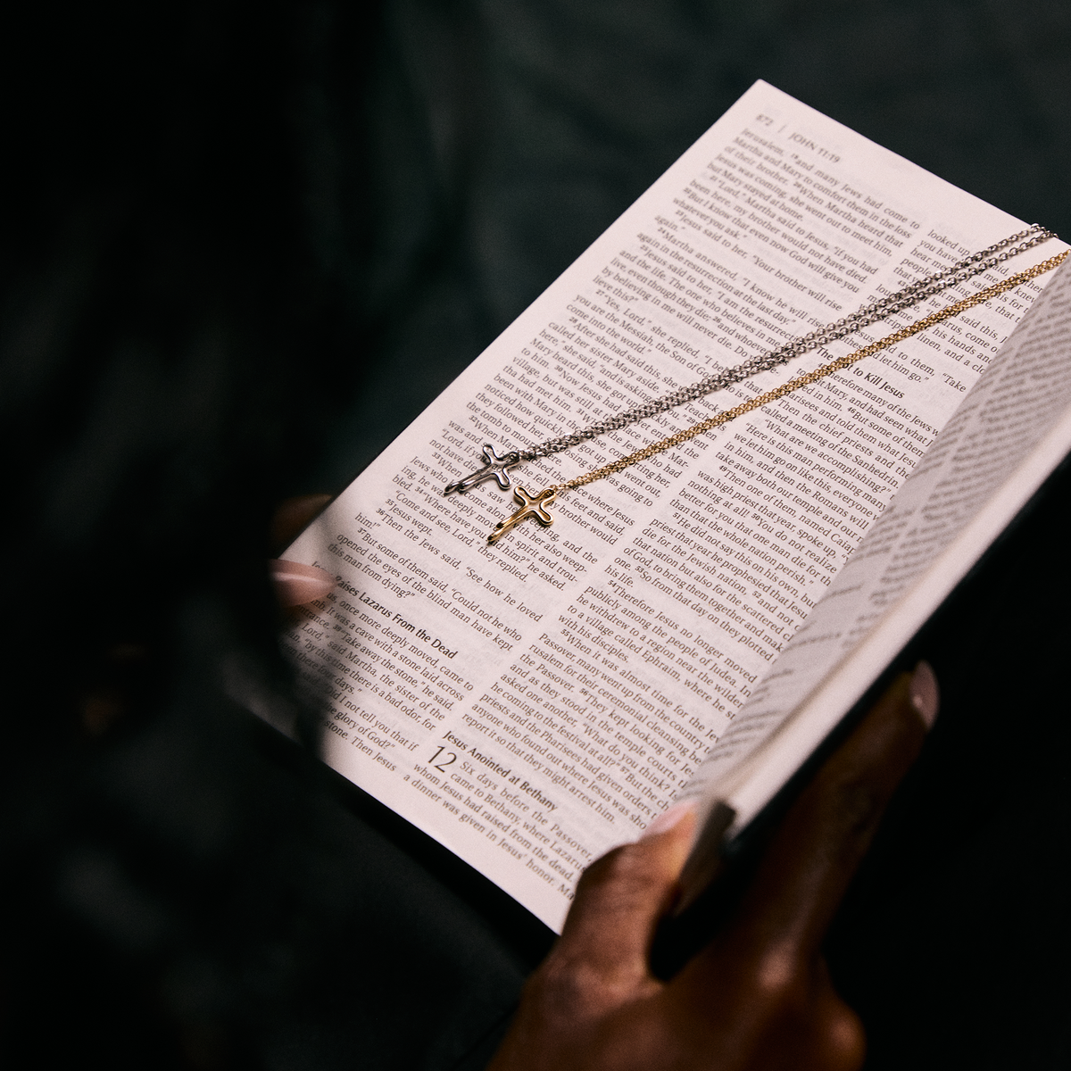 Two cross pendants on chains draped over an open Bible, held by hands in a dark setting.