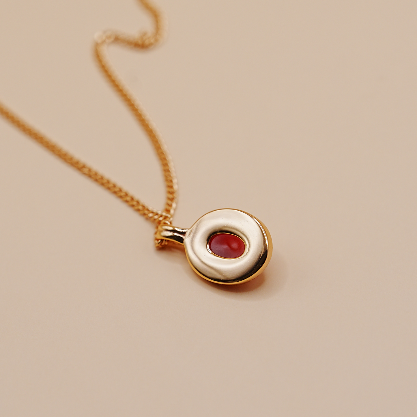 A gold chain necklace with a circular pendant featuring a polished gold outer ring and a central oval red gemstone, displayed on a solid beige background.