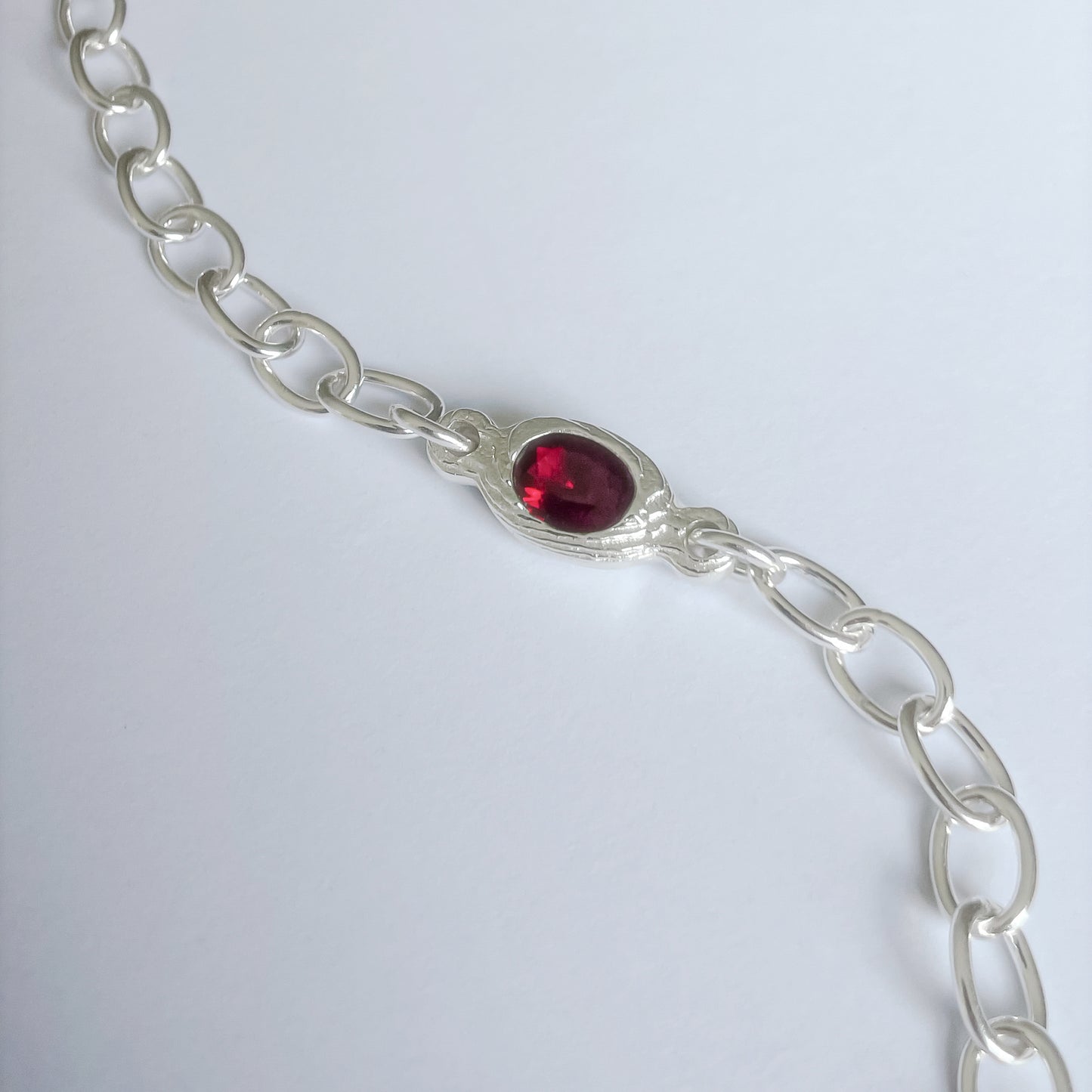 A silver chain bracelet featuring a central oval red gemstone in a textured bezel setting, photographed on a light gray background.