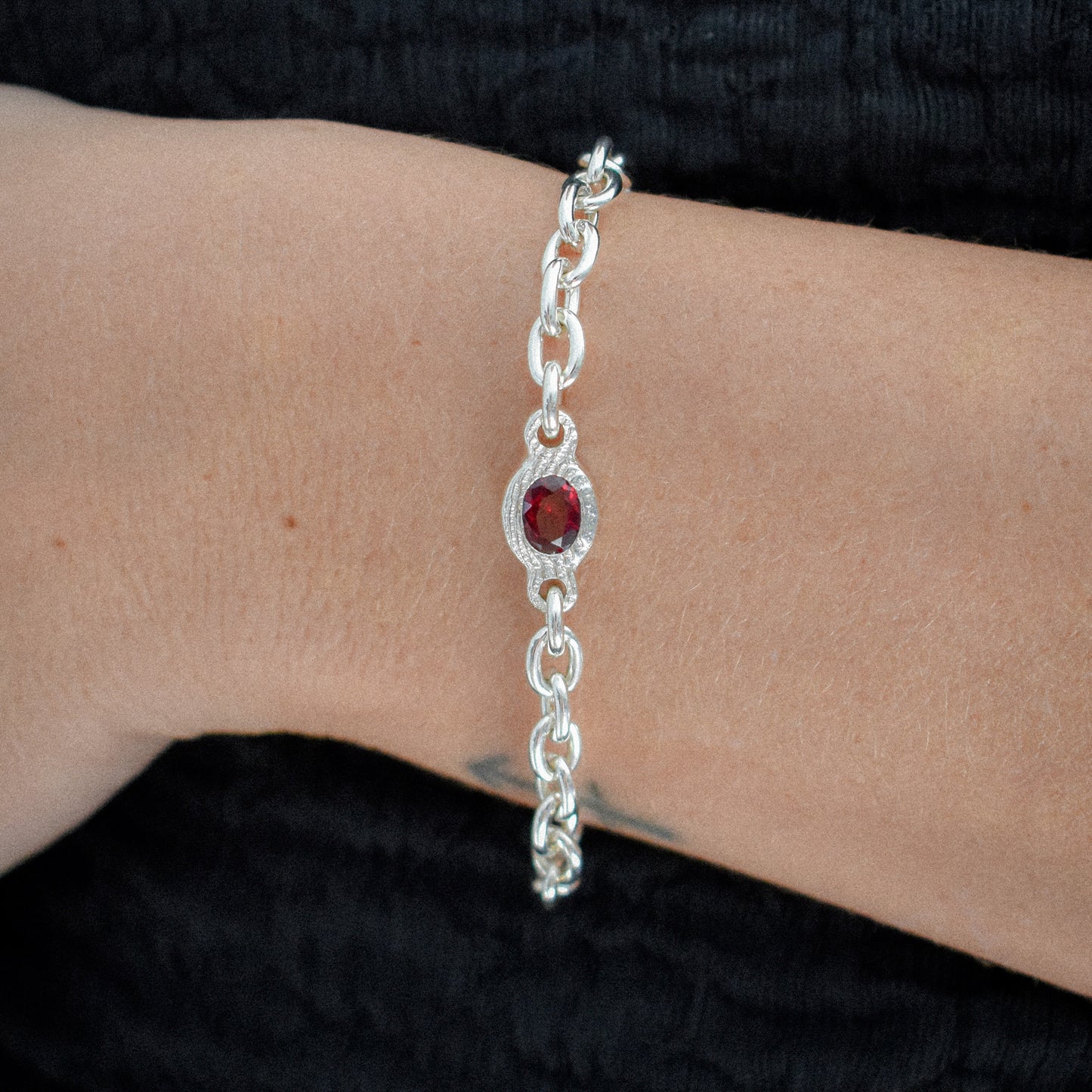 A sterling silver chain bracelet with a central oval red gemstone surrounded by small clear stones, shown worn on a person's wrist against a black textured background.