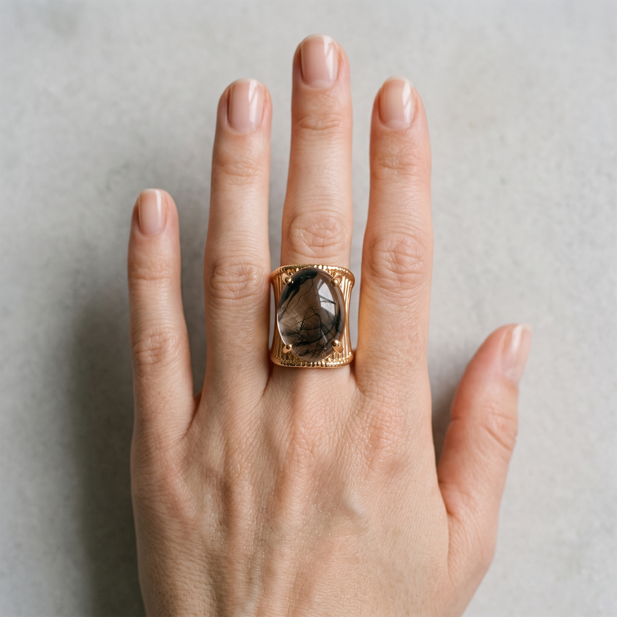 A close-up of a hand wearing a large gold signet ring with a rectangular smoky gray gemstone, set against a neutral gray background.