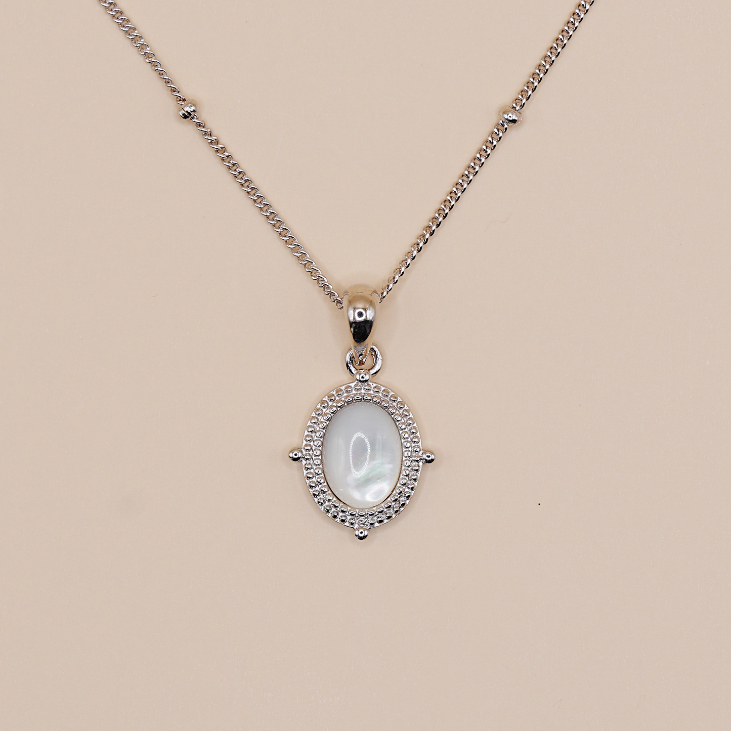 A silver chain necklace with an oval pendant featuring a white mother-of-pearl center stone surrounded by a halo of small clear stones, displayed on a solid beige background.