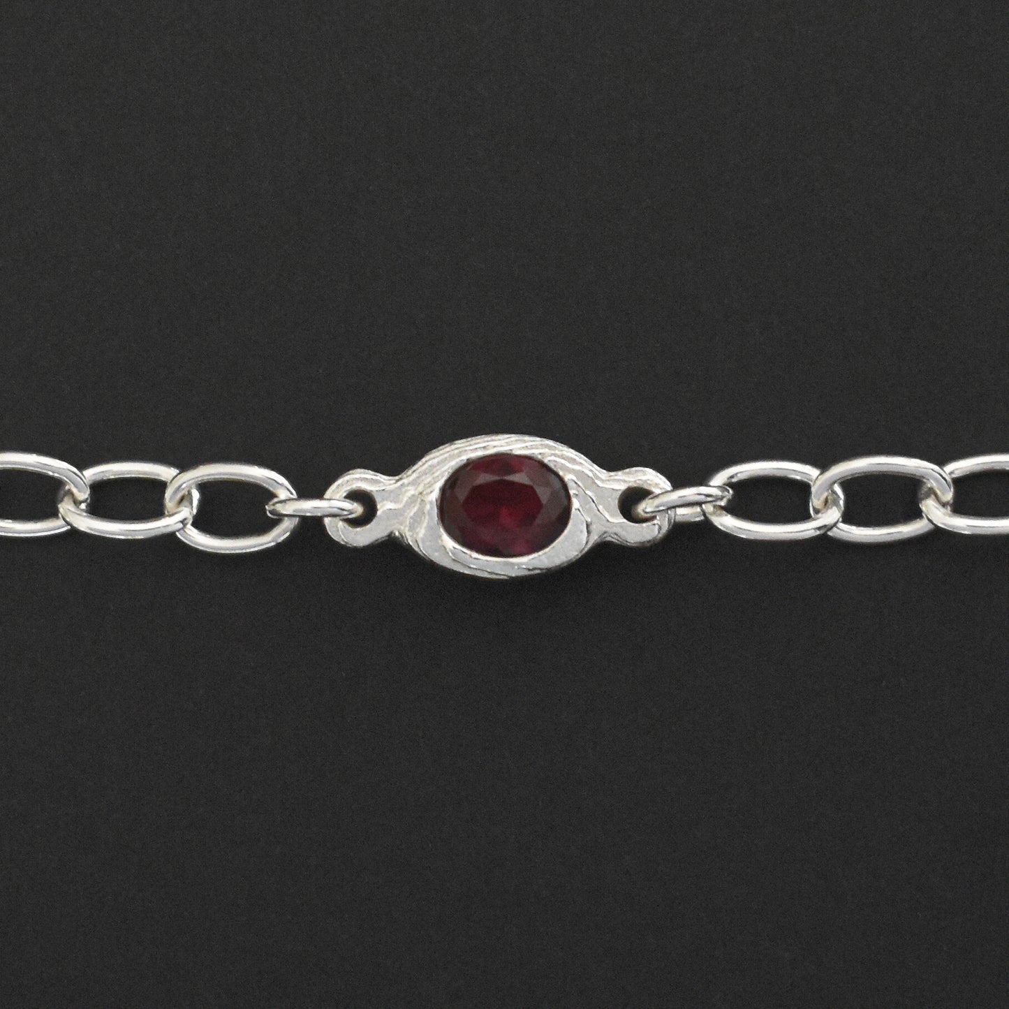 A sterling silver chain bracelet featuring a central oval bezel-set dark red gemstone, shown against a solid black background.
