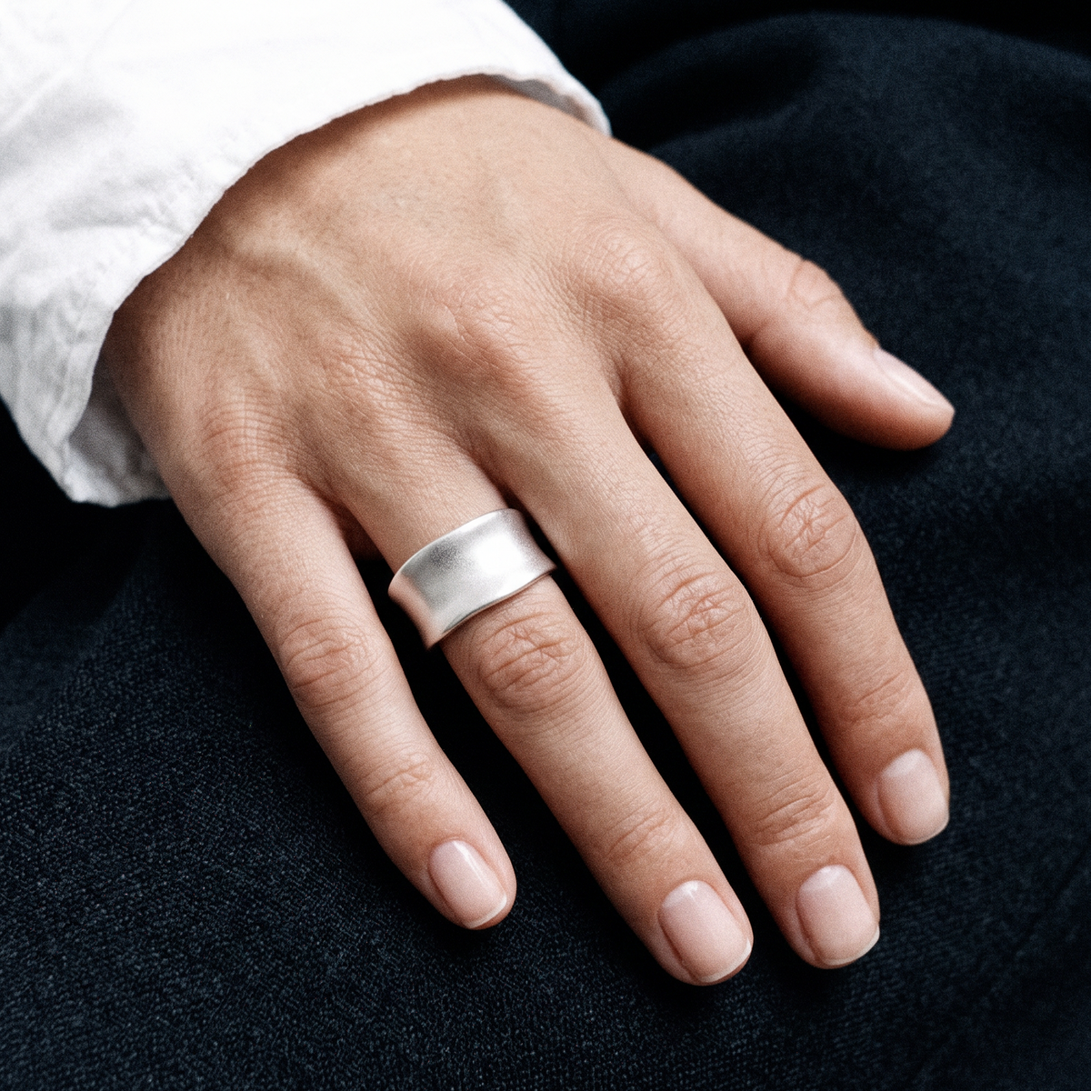 A close-up of a person's hand wearing a wide, polished silver band ring, resting on dark navy fabric.