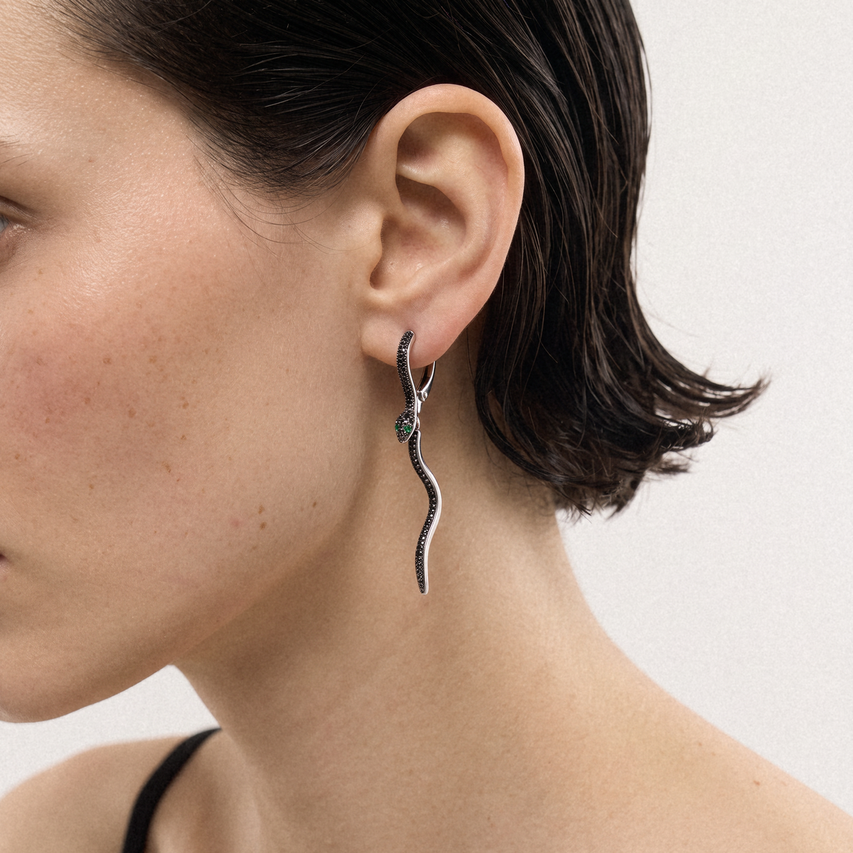 A close-up of a model's ear wearing a silver wavy dangle earring adorned with small black stones, set against a plain light background.