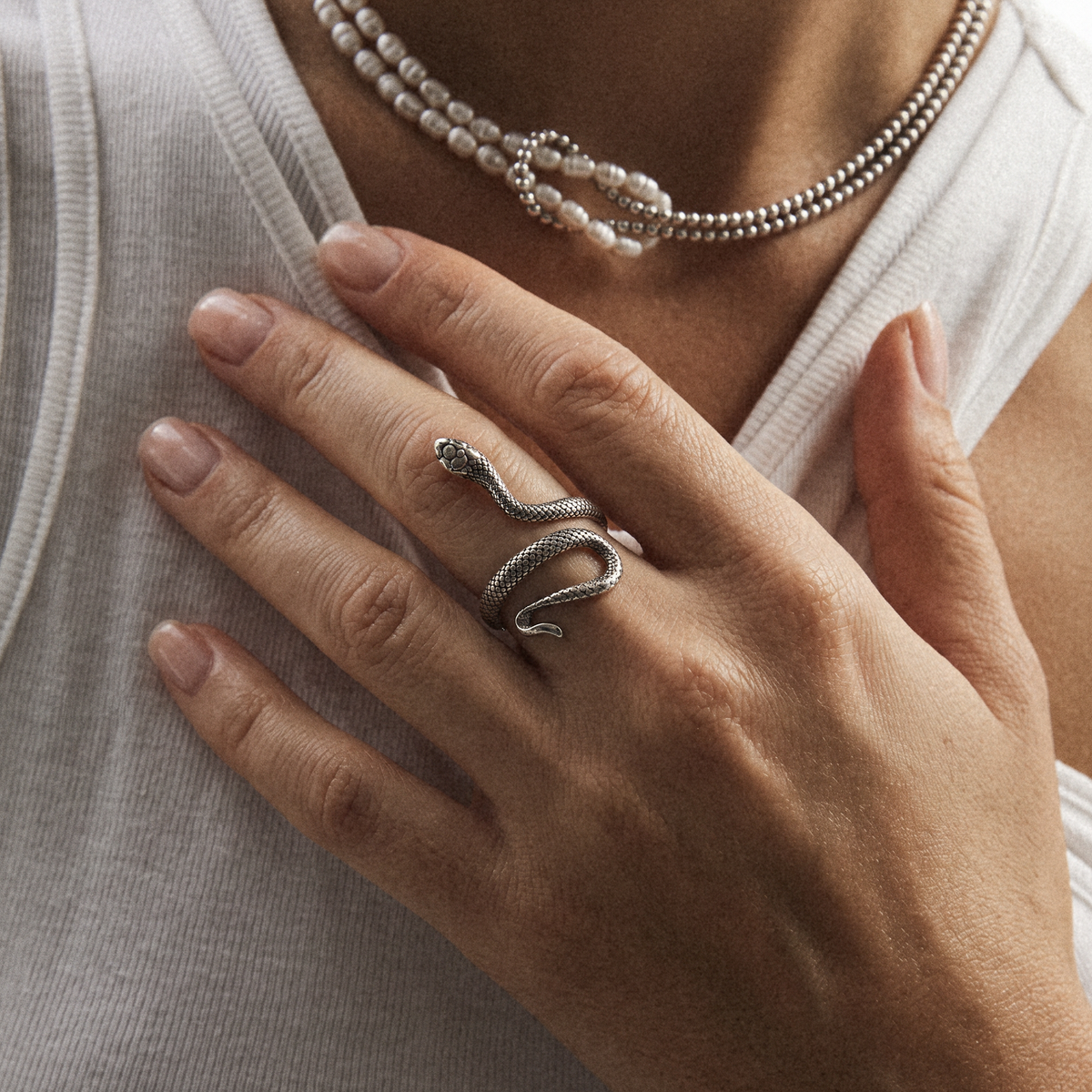 A close-up of a person's hand wearing a silver snake ring, styled with a pearl beaded necklace and a white ribbed top.