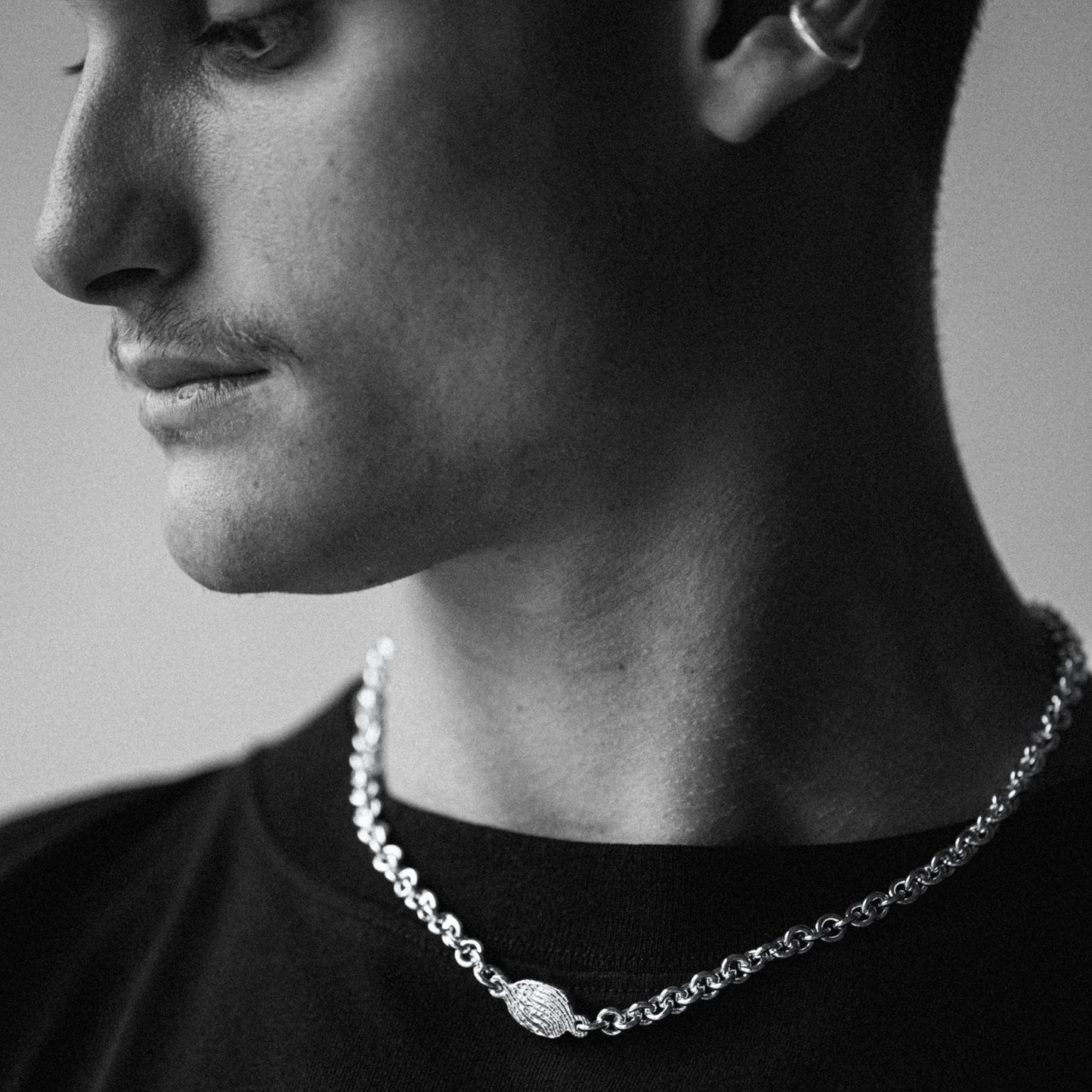A close-up black and white photo of a man's profile wearing a silver chain necklace with a textured oval pendant.