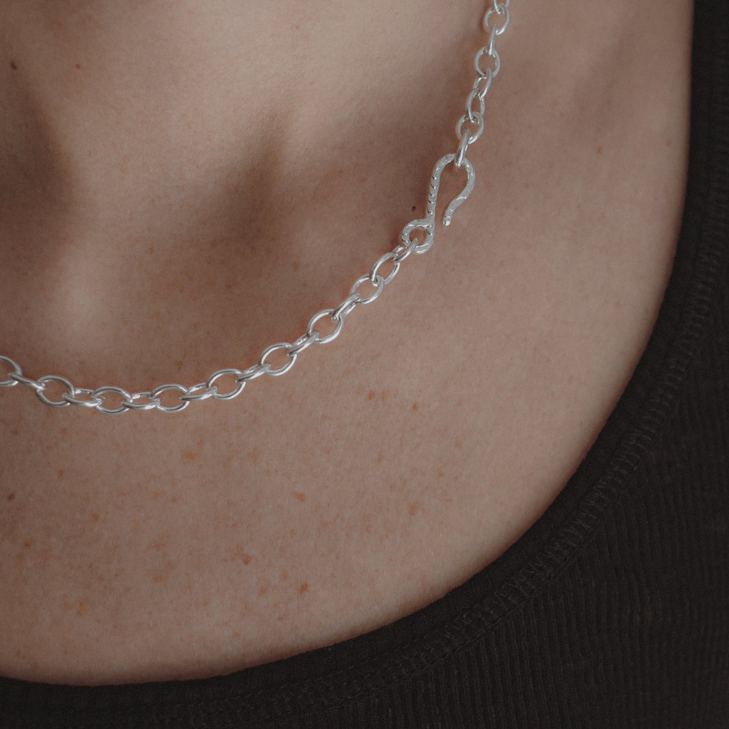 A close-up of a delicate silver chain necklace worn on a person's neck, featuring a small heart-shaped pendant and oval links, with a black ribbed top visible below.