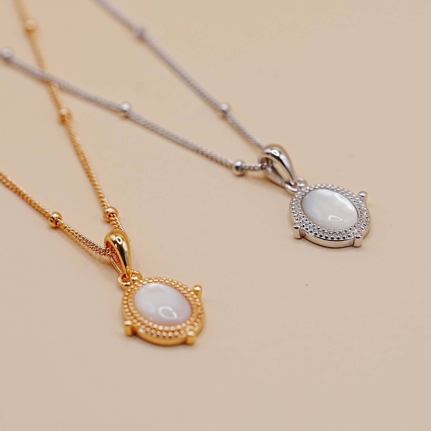 Two oval pendant necklaces on delicate chains, one in gold and one in silver, featuring milky white oval cabochon stones surrounded by small pavé-set clear stones, displayed on a warm beige background.