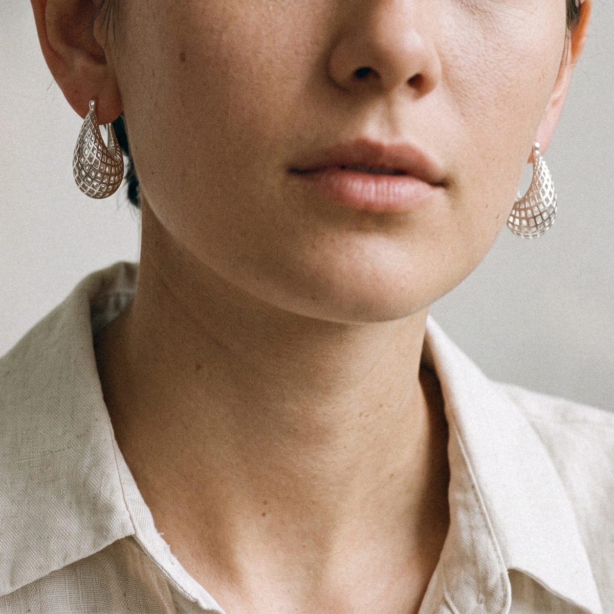 Close-up of a person wearing textured silver hoop earrings and a light beige collared shirt against a soft neutral background.