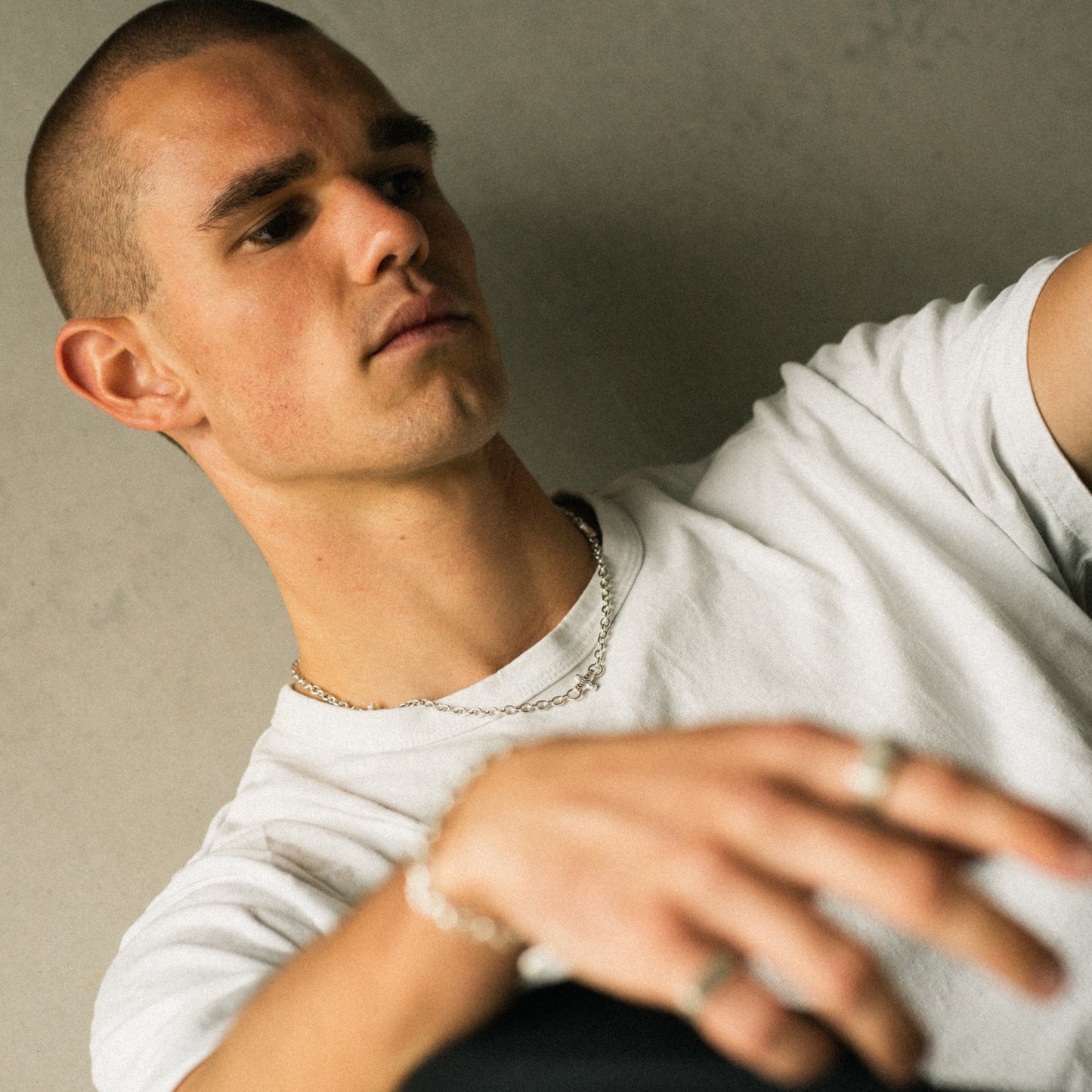 A close-up lifestyle shot of a man wearing a white crewneck t-shirt, silver chain necklace, and silver rings, leaning against a textured gray wall.