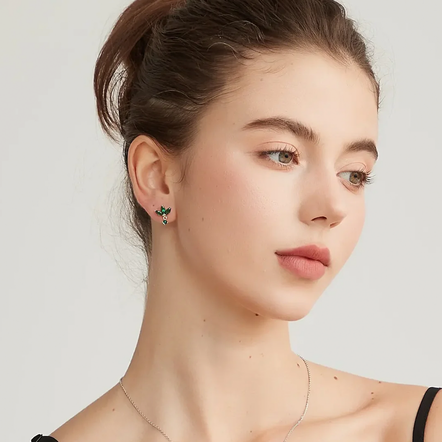 A close-up of a woman wearing a small green and gold butterfly stud earring, with her hair in a bun and a delicate silver necklace visible against a light gray background.
