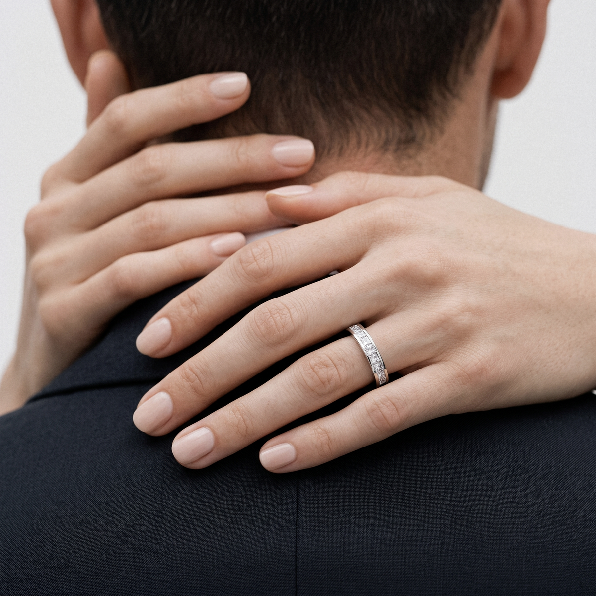A close-up lifestyle shot of a woman's hands resting on a man's neck and shoulder, wearing a slim silver band ring with small clear stones on her ring finger.