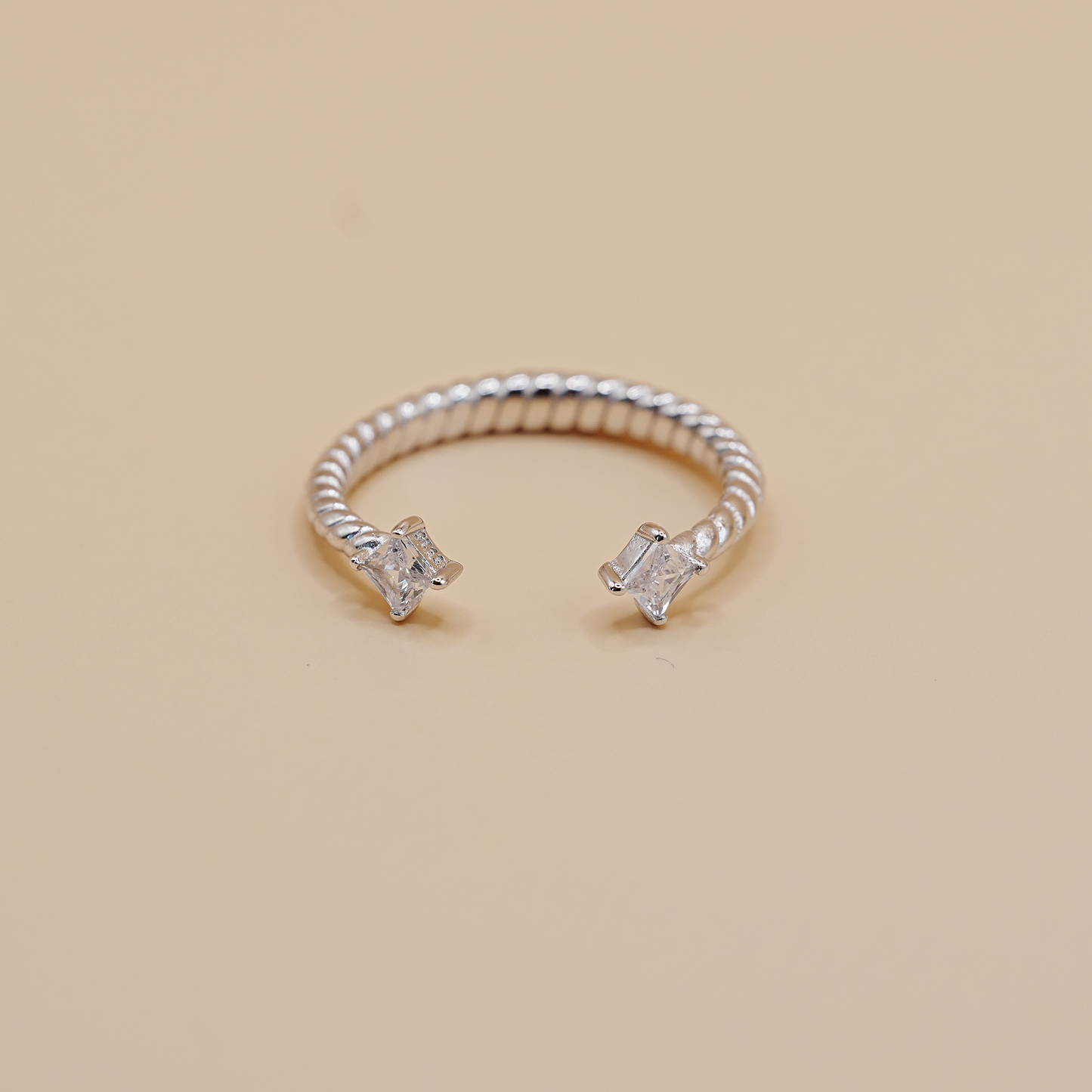 A silver twisted rope open ring with two clear baguette and round-cut stones, displayed on a solid beige background.