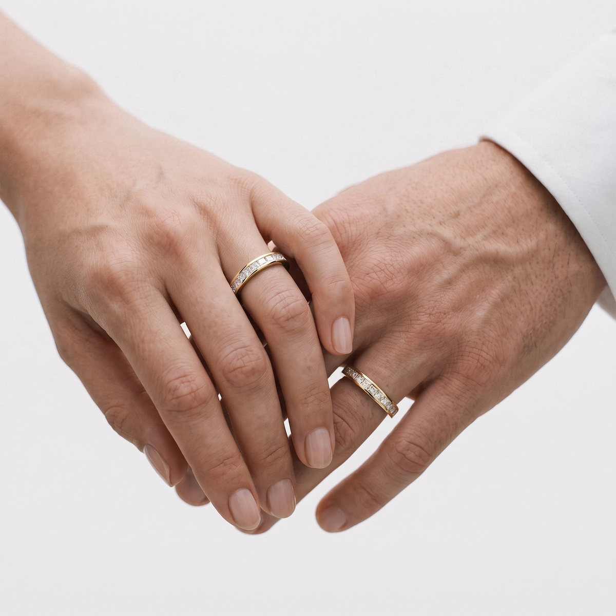 Two hands gently touching, each wearing a slim silver band ring with small clear stones, photographed against a soft light gray background.