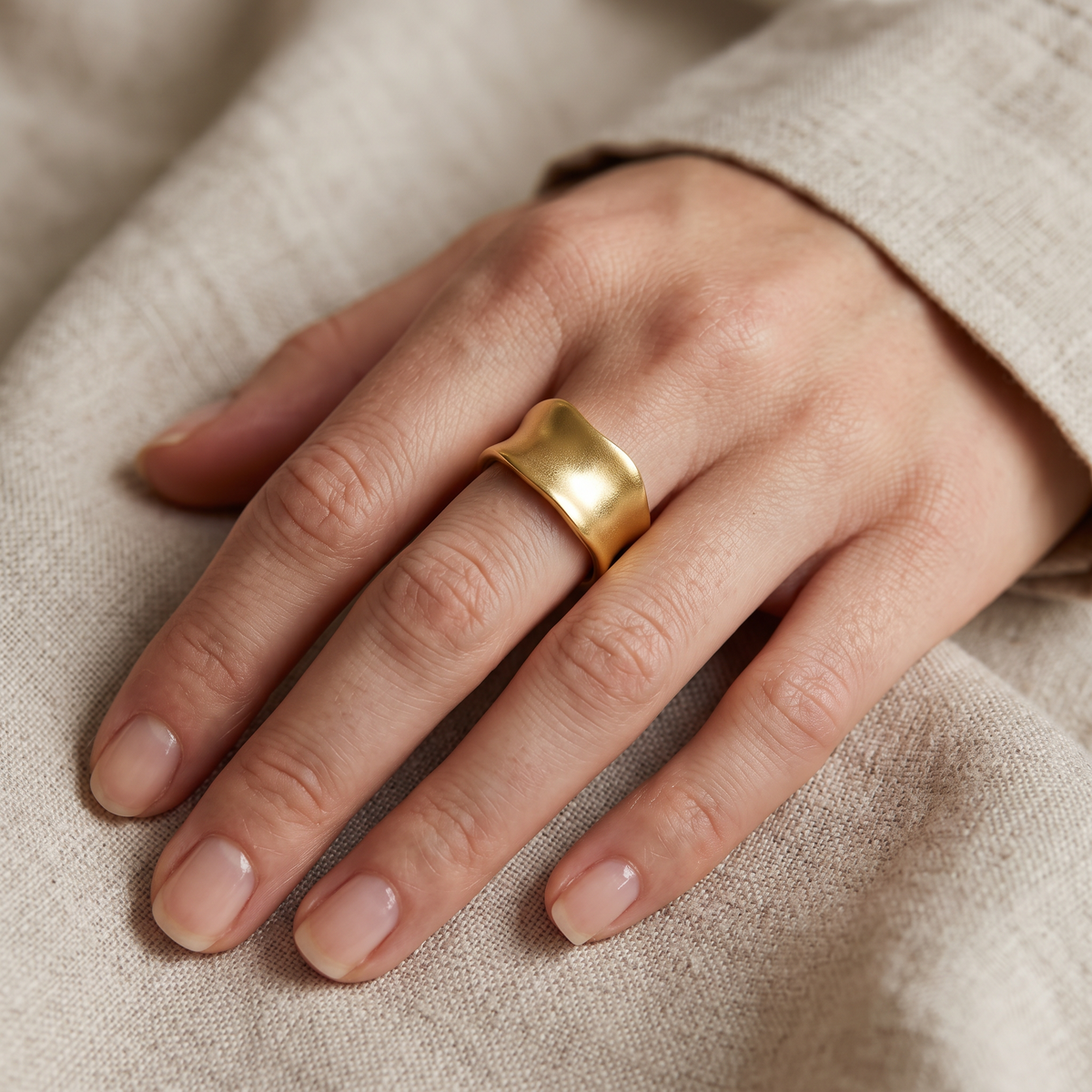 A close-up of a hand wearing a wide, polished gold ring, resting on a textured beige fabric background.