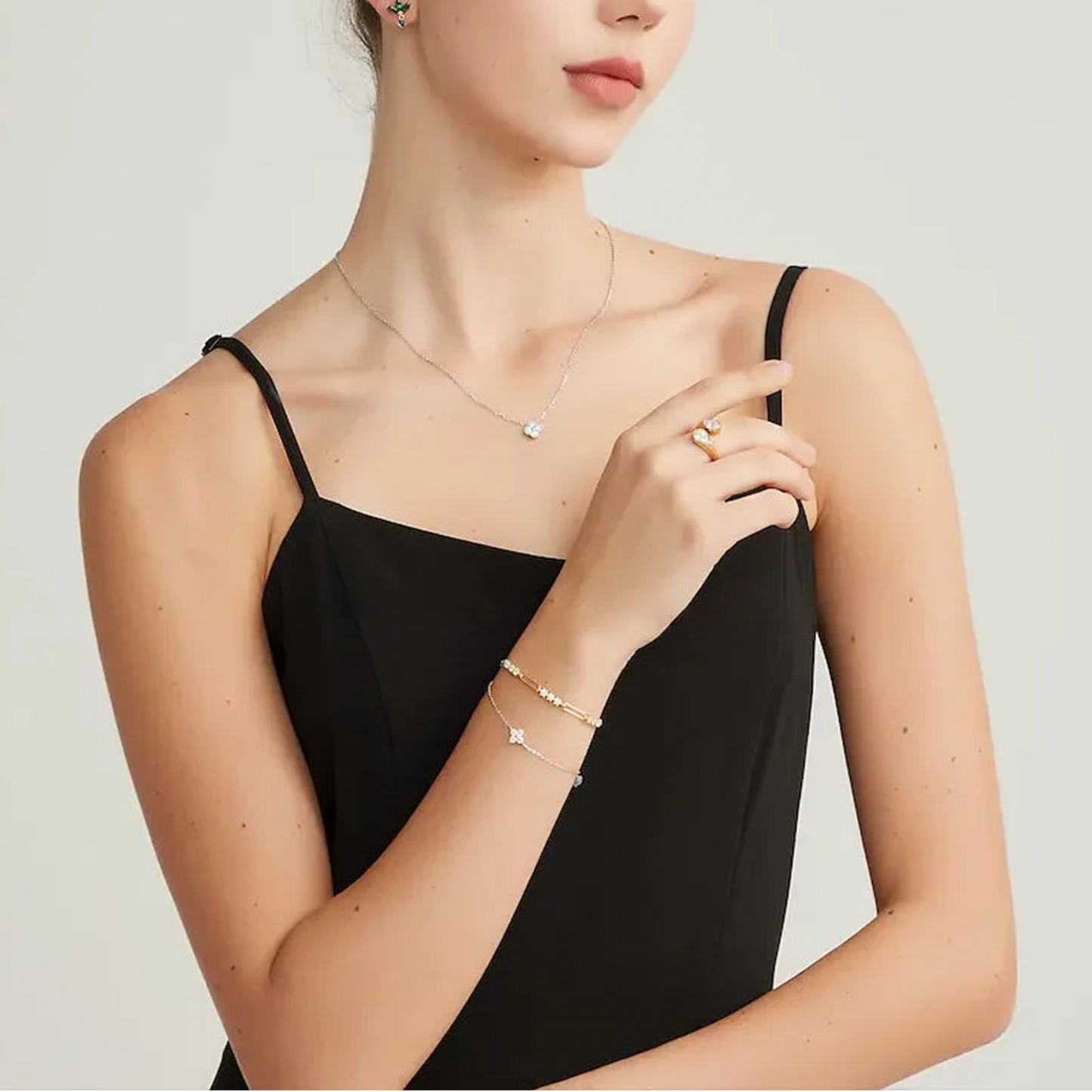 A woman wearing delicate jewelry including a thin silver necklace, a gold ring, and two gold bracelets with star details, styled with a black camisole against a light gray background.