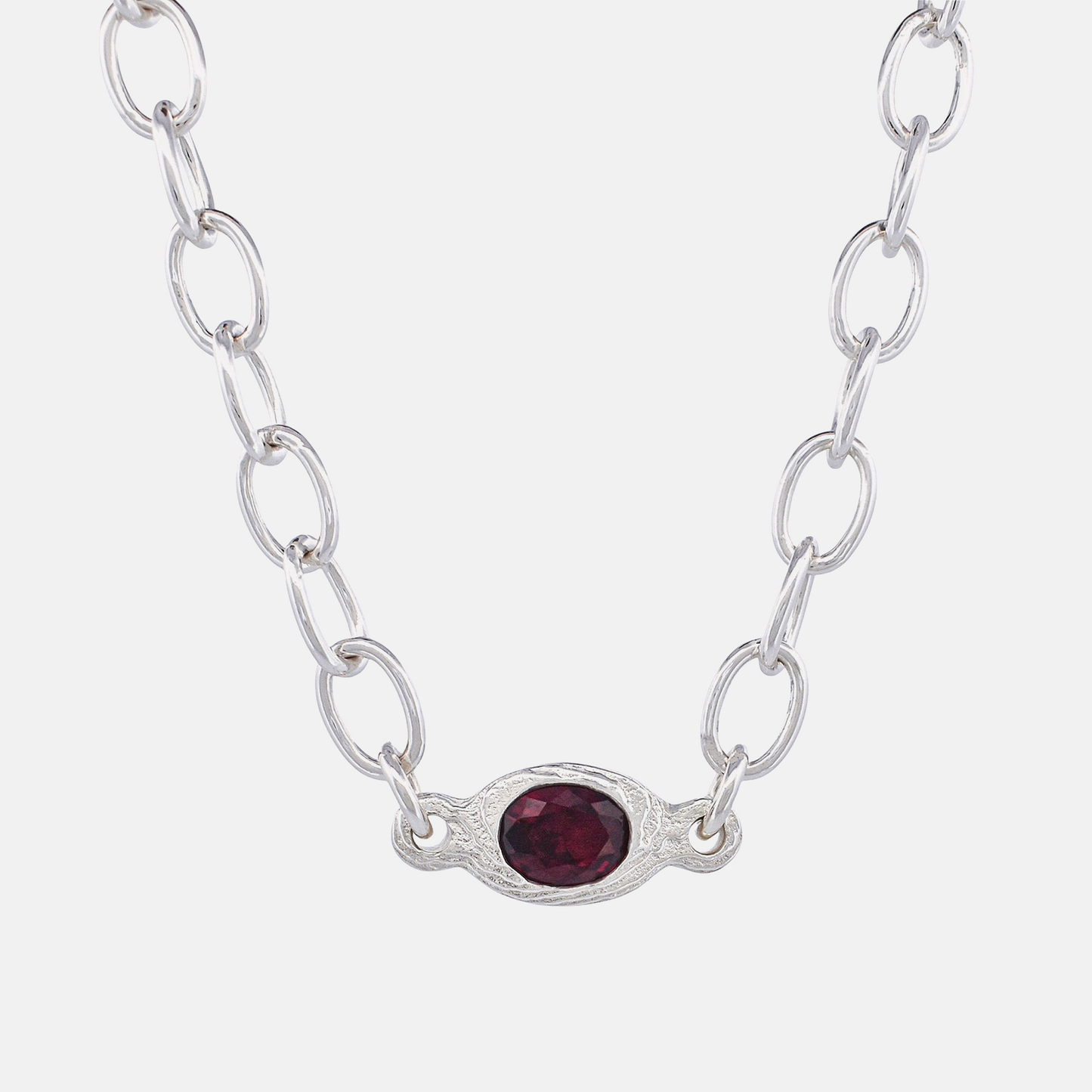 A silver link chain necklace featuring an oval silver pendant with a textured border and a faceted dark red gemstone in the center, set against a plain light gray background.