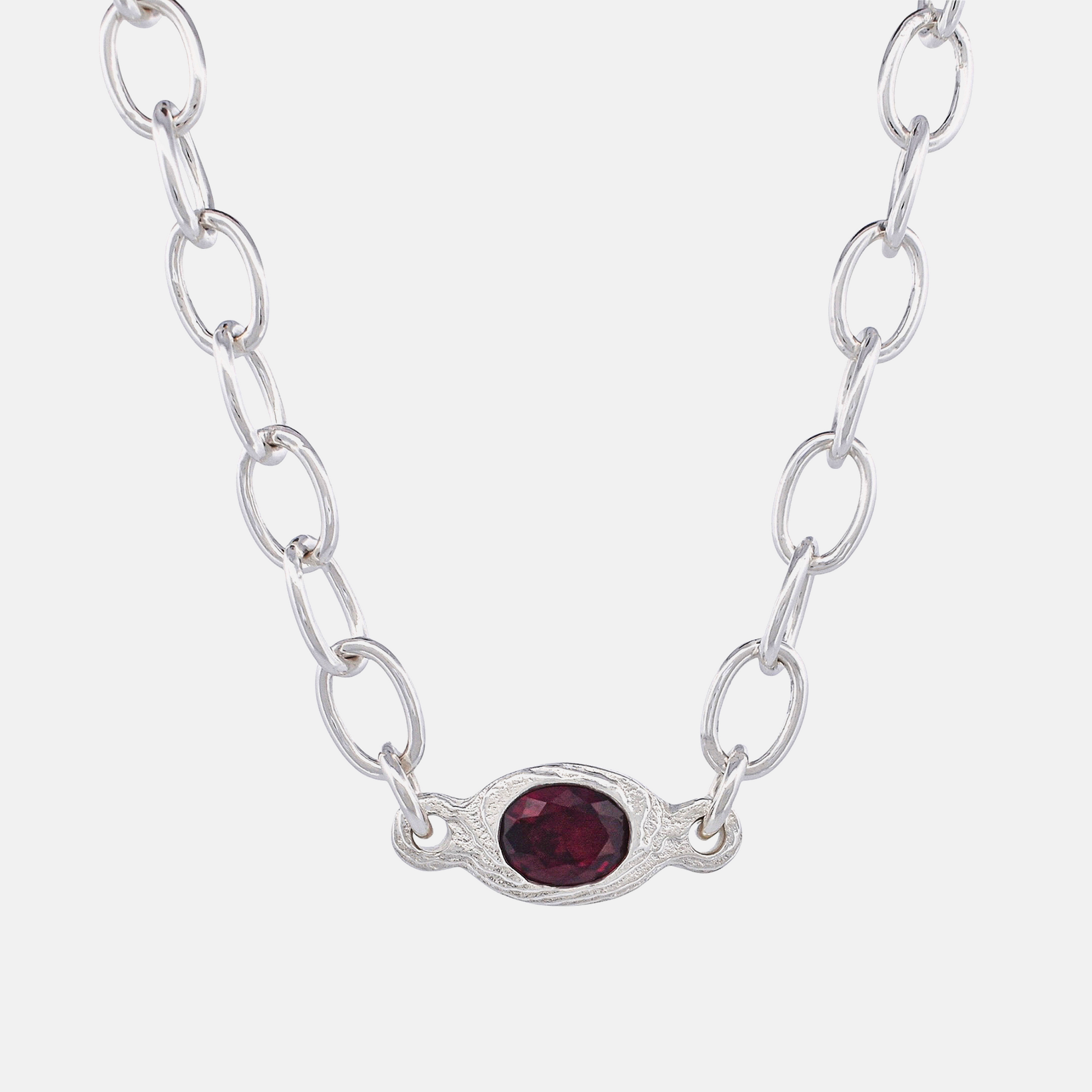 A silver link chain necklace featuring an oval silver pendant with a textured border and a faceted dark red gemstone in the center, set against a plain light gray background.