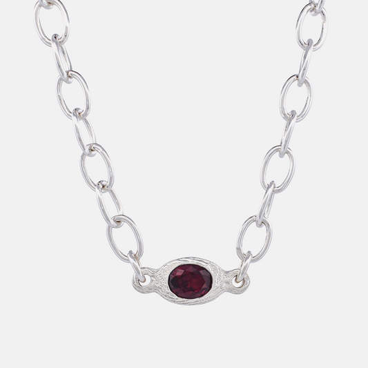 A silver link chain necklace featuring an oval silver pendant with a textured border and a faceted dark red gemstone in the center, set against a plain light gray background.