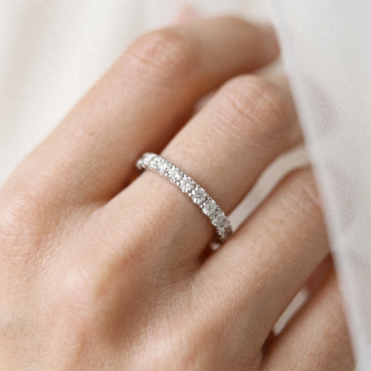 A close-up of a hand wearing a thin silver eternity ring set with small clear stones, photographed against a soft white fabric background.