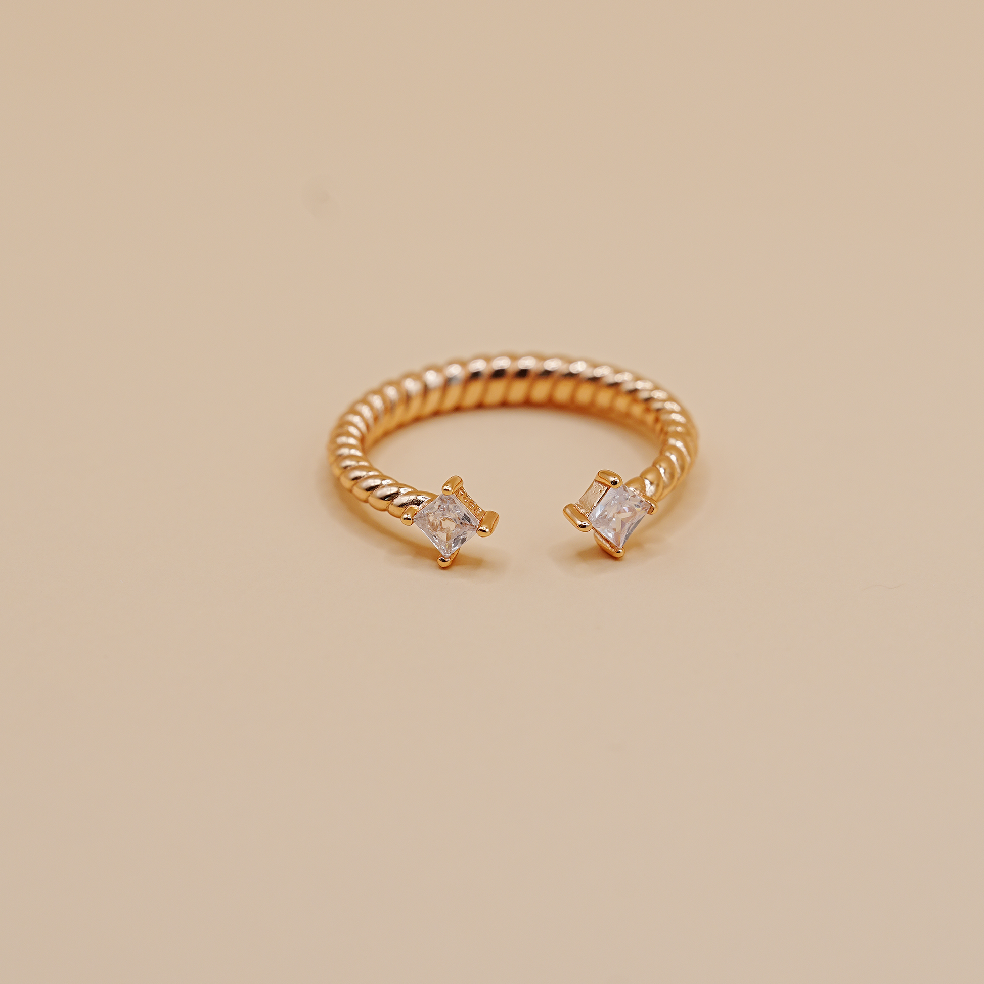A gold-toned twisted rope open ring with two clear rectangular gemstones, photographed on a plain warm beige background.