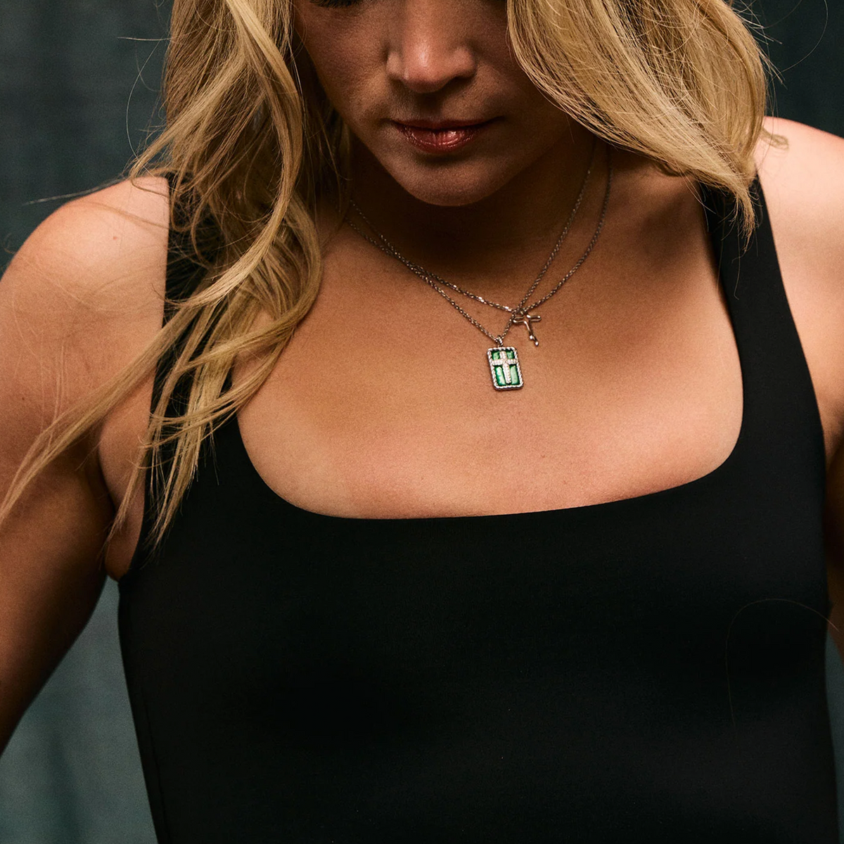 A close-up of a woman wearing a silver-toned double-strand necklace with a small cross and a rectangular pendant featuring a green and white cross design, paired with a black tank top.