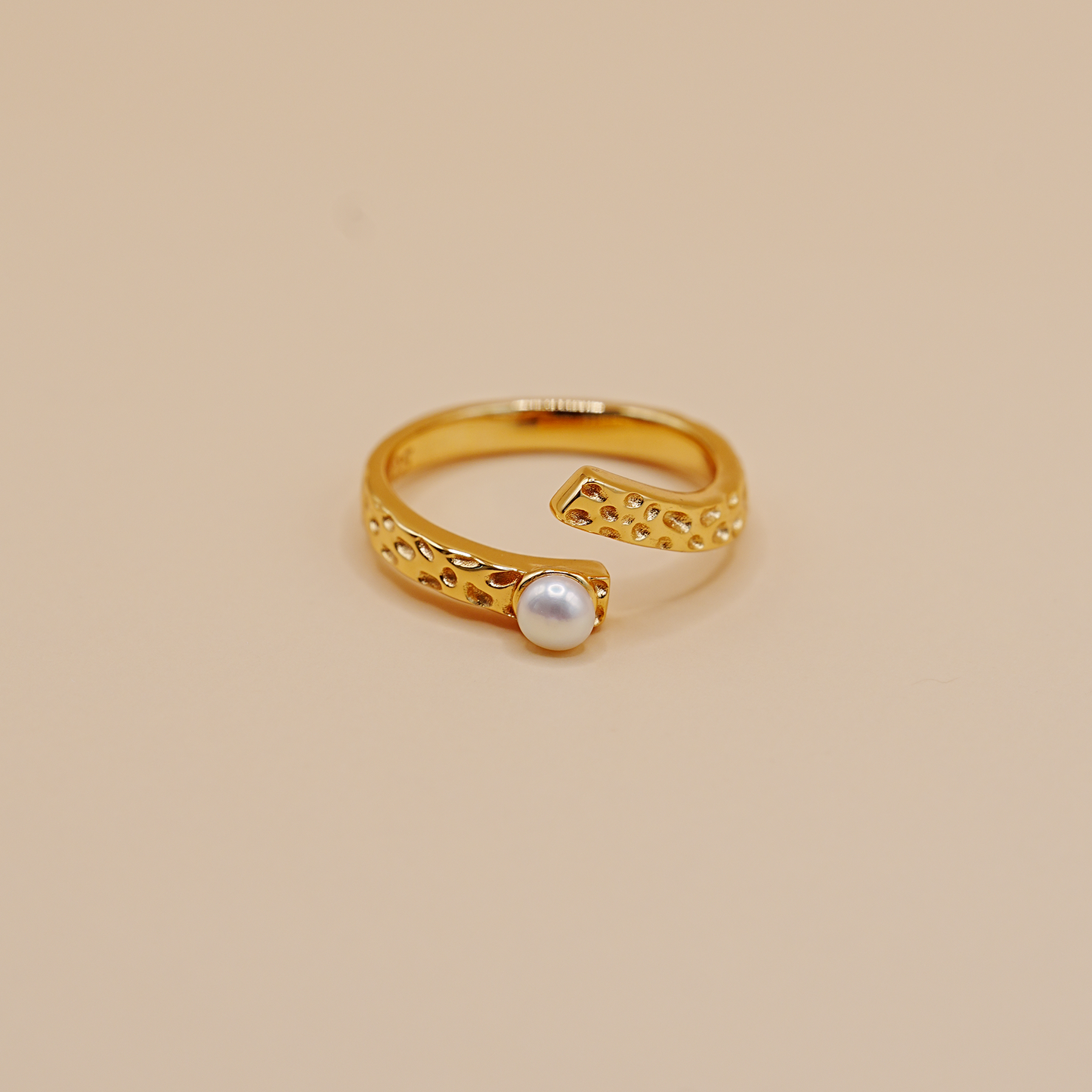 A gold open-band ring with a textured, perforated design and a single round white pearl set in the center, photographed on a solid beige background.