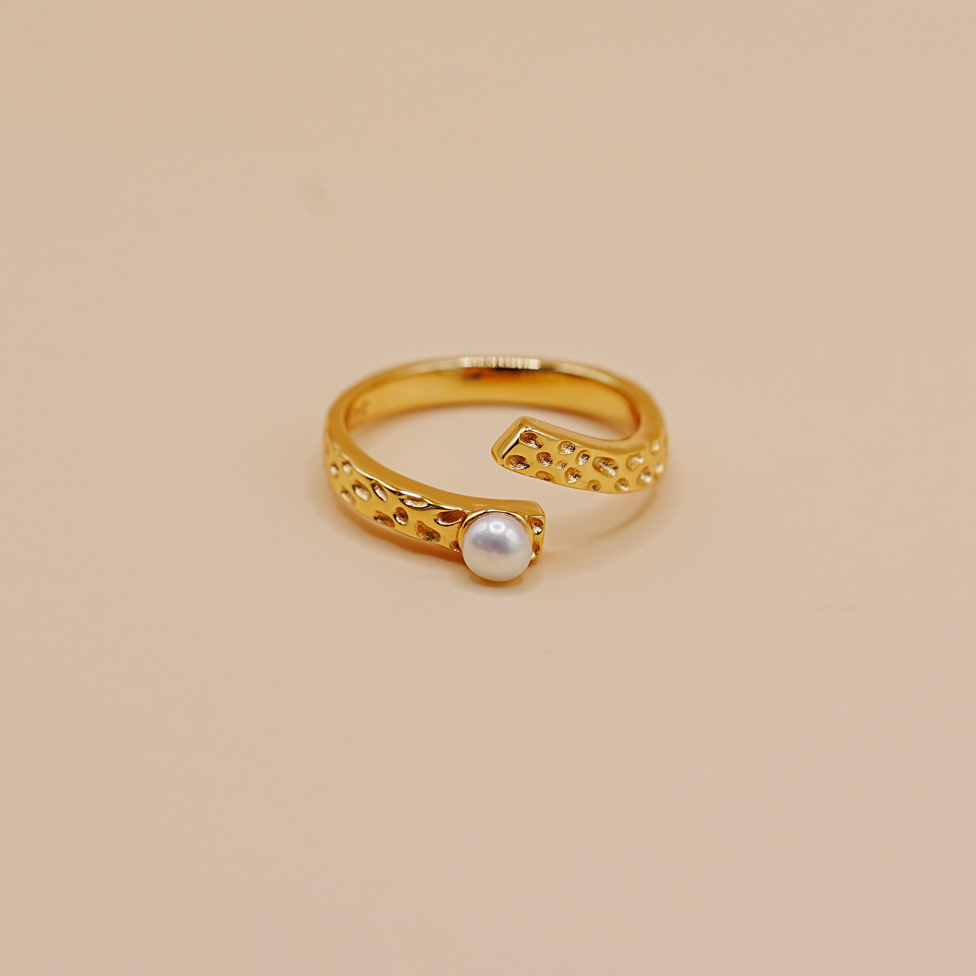A gold open-band ring with a textured, perforated design and a single round white pearl set in the center, photographed on a solid beige background.
