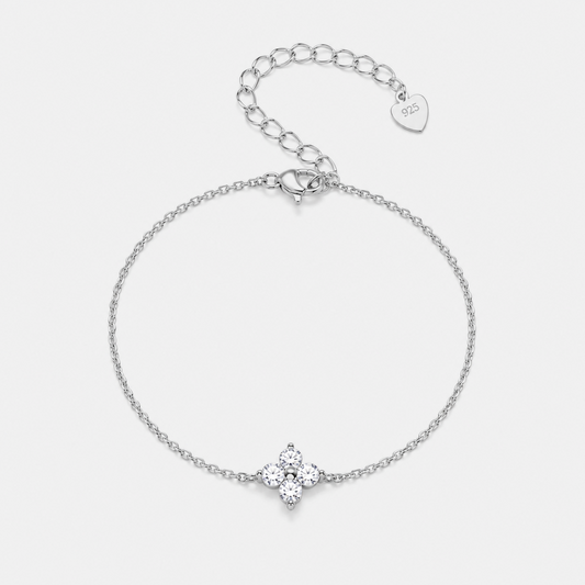 A delicate silver chain bracelet with a small four-petal flower charm set with clear stones, shown on a light gray background. The bracelet includes an adjustable link chain extender with a heart-shaped tag engraved with '14K'.