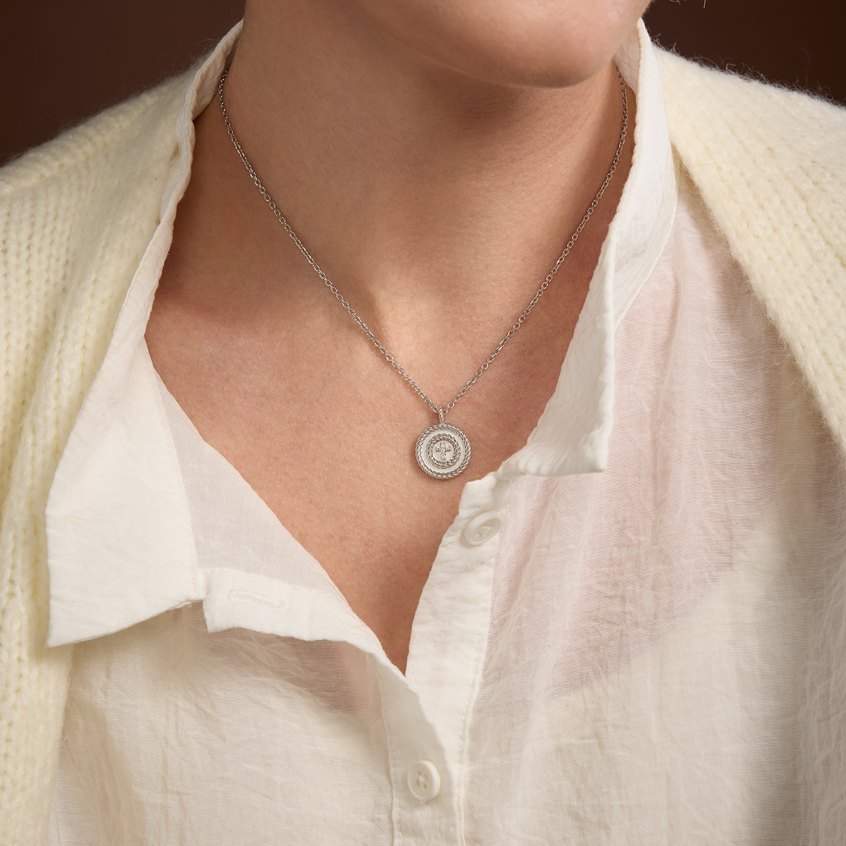 A close-up of a person wearing a delicate silver chain cross necklace with a circular pendant, styled over a white collared shirt and cream cardigan.