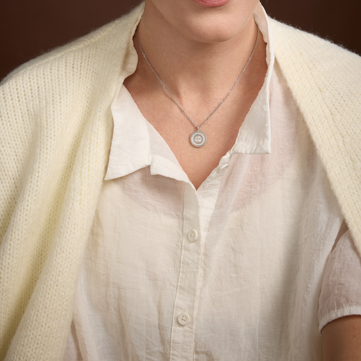 A close-up of a person wearing a delicate silver cross necklace with a circular pendant, styled over a cream-colored linen shirt and a matching knit cardigan.