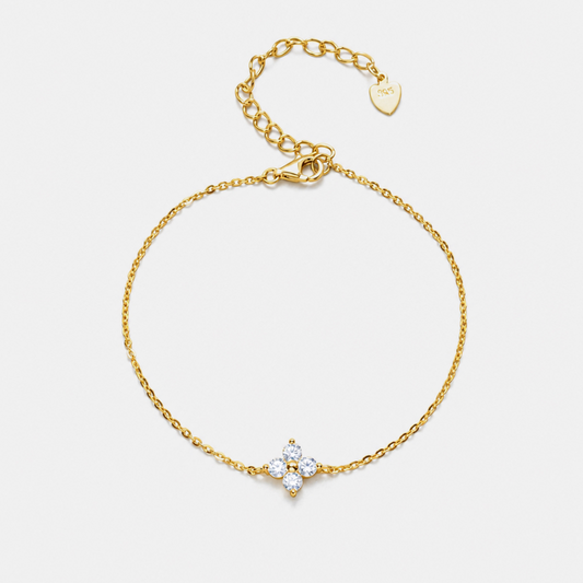 A delicate gold chain bracelet with a small four-petal flower charm set with clear stones, displayed on a light gray background.