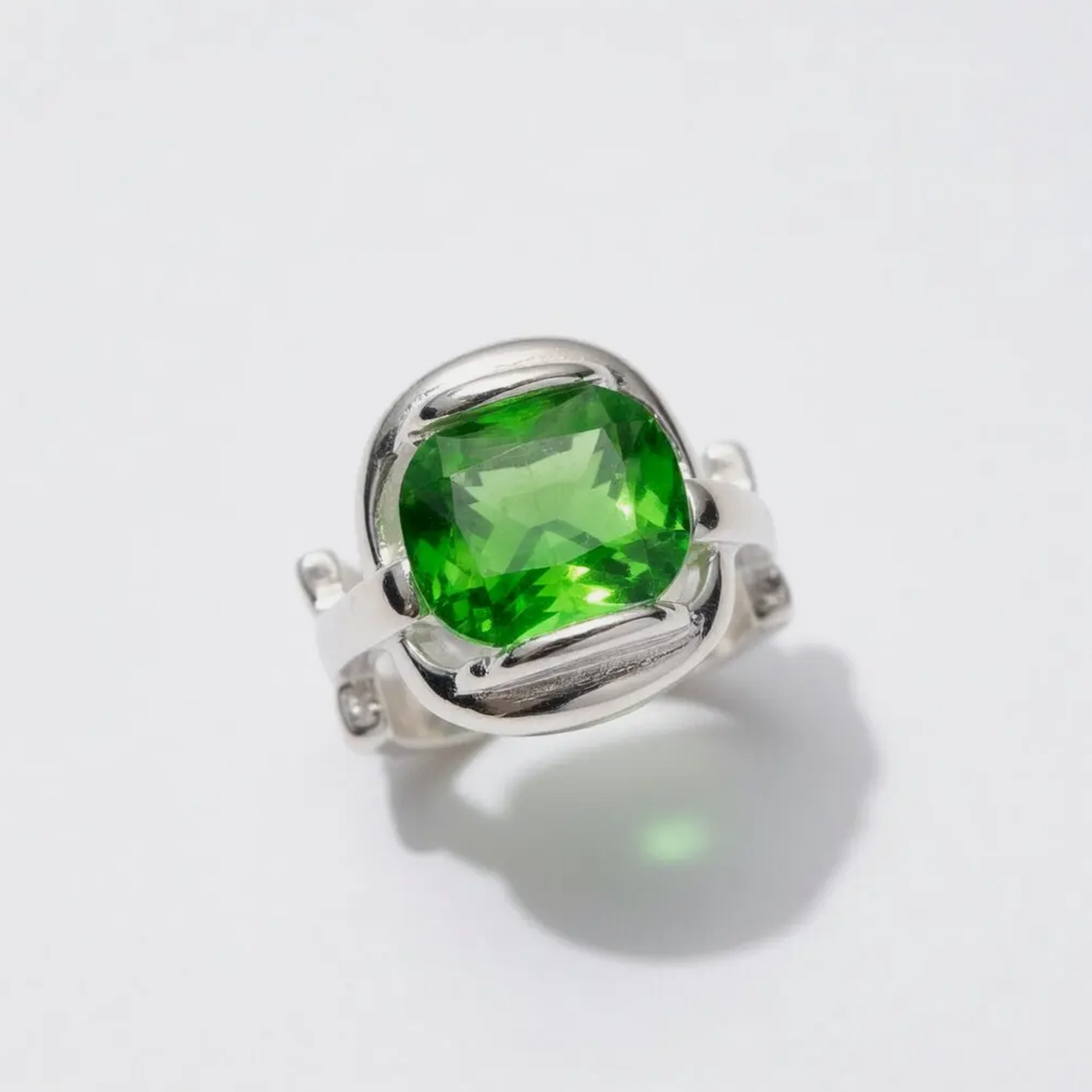 A silver ring featuring a large, cushion-cut green gemstone in a prong setting, photographed on a clean white background with a soft shadow.
