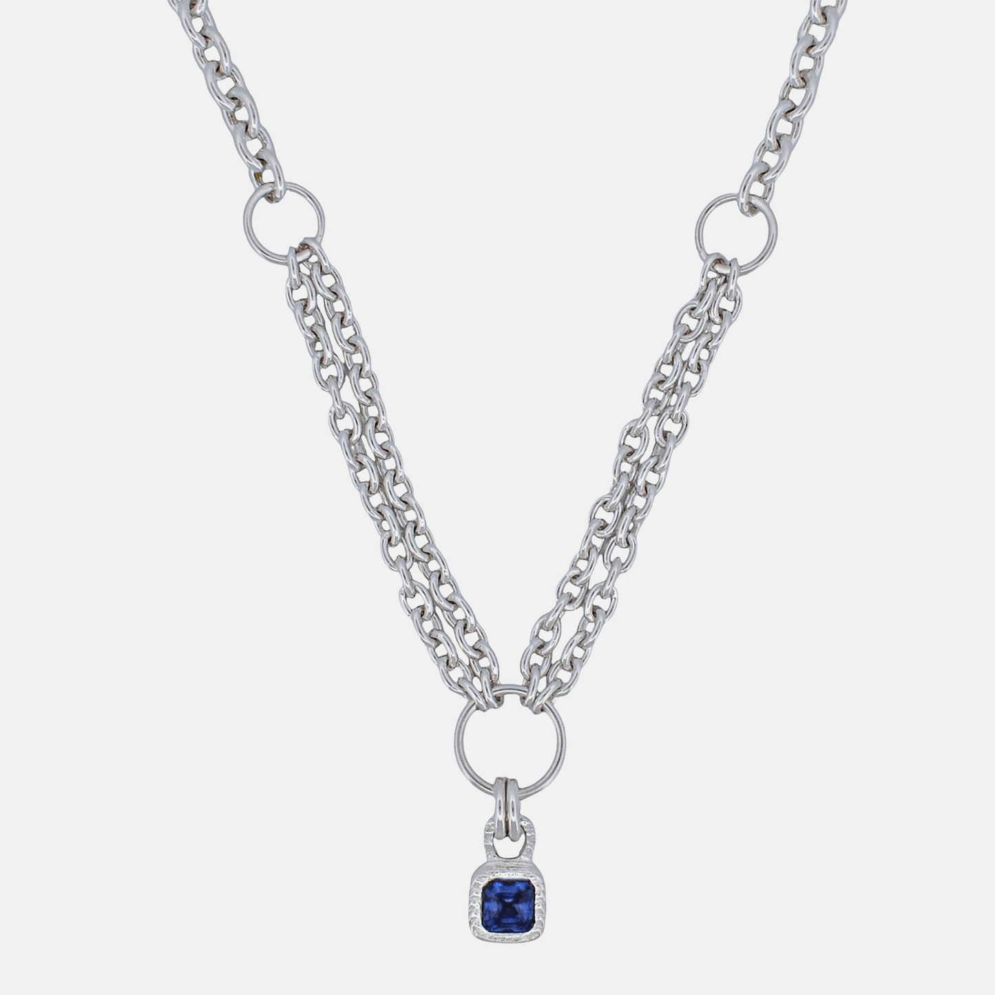 A silver-tone double-strand chain necklace featuring a central ring and a small square pendant with a deep blue gemstone surrounded by a clear crystal border, set against a light gray background.