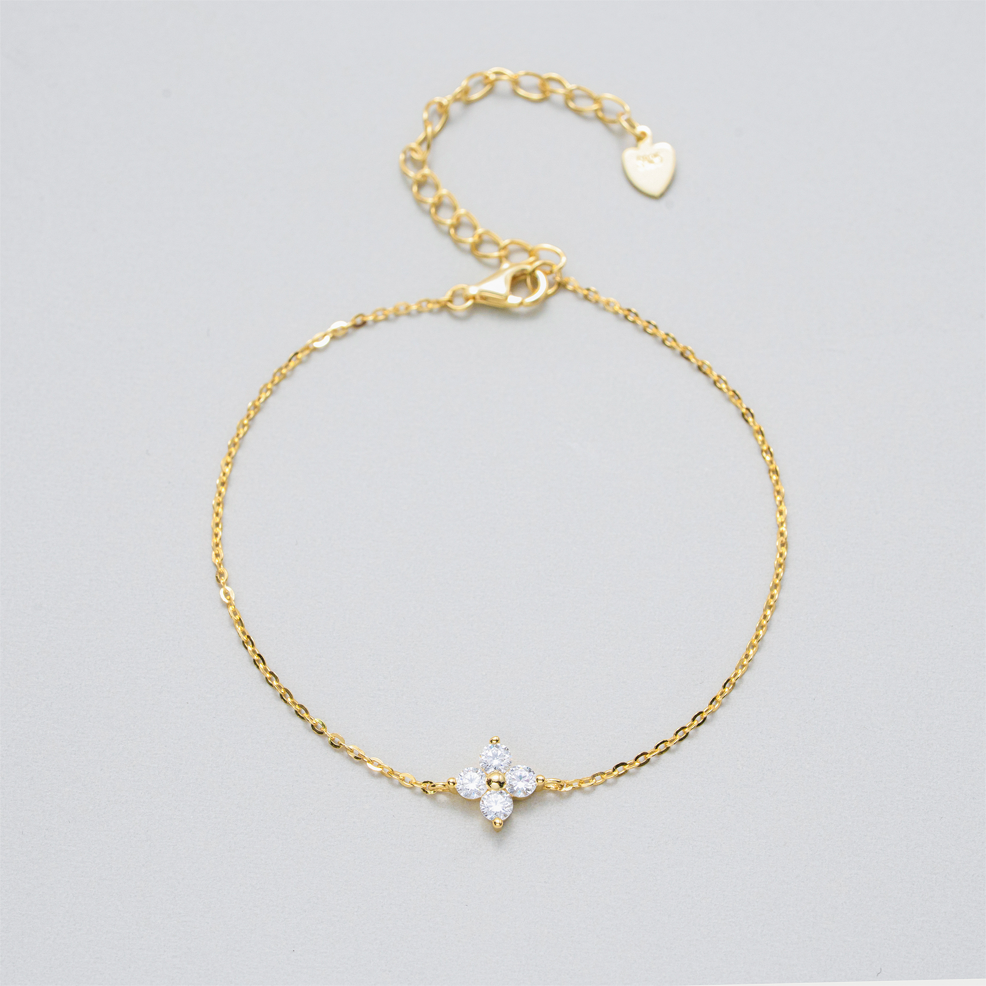 A delicate gold chain bracelet with a small flower-shaped charm set with clear stones, displayed on a light gray background.