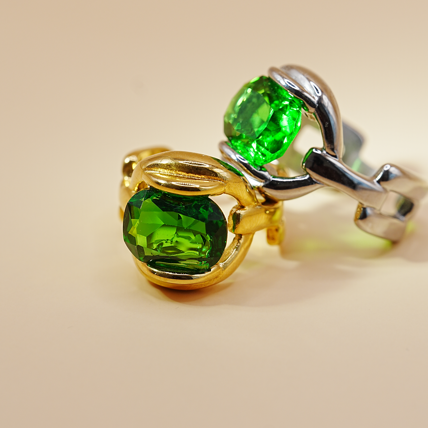 A close-up studio shot of a ring with two oval-cut vivid green gemstones in gold and silver settings, arranged on a warm beige background.