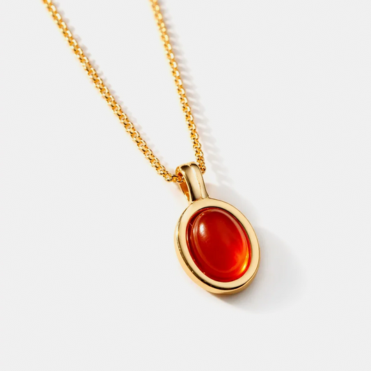 A gold chain necklace with an oval pendant featuring a smooth, polished red cabochon gemstone, set against a clean light gray background.